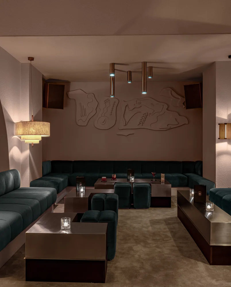 Modern lounge area with dark green velvet sofas, low metal coffee tables with candles, ambient pendant and wall lighting, and abstract wall art.