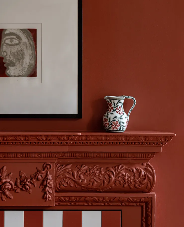 Decorative white pitcher with red floral patterns placed on an ornate red fireplace mantel next to a framed abstract face artwork.