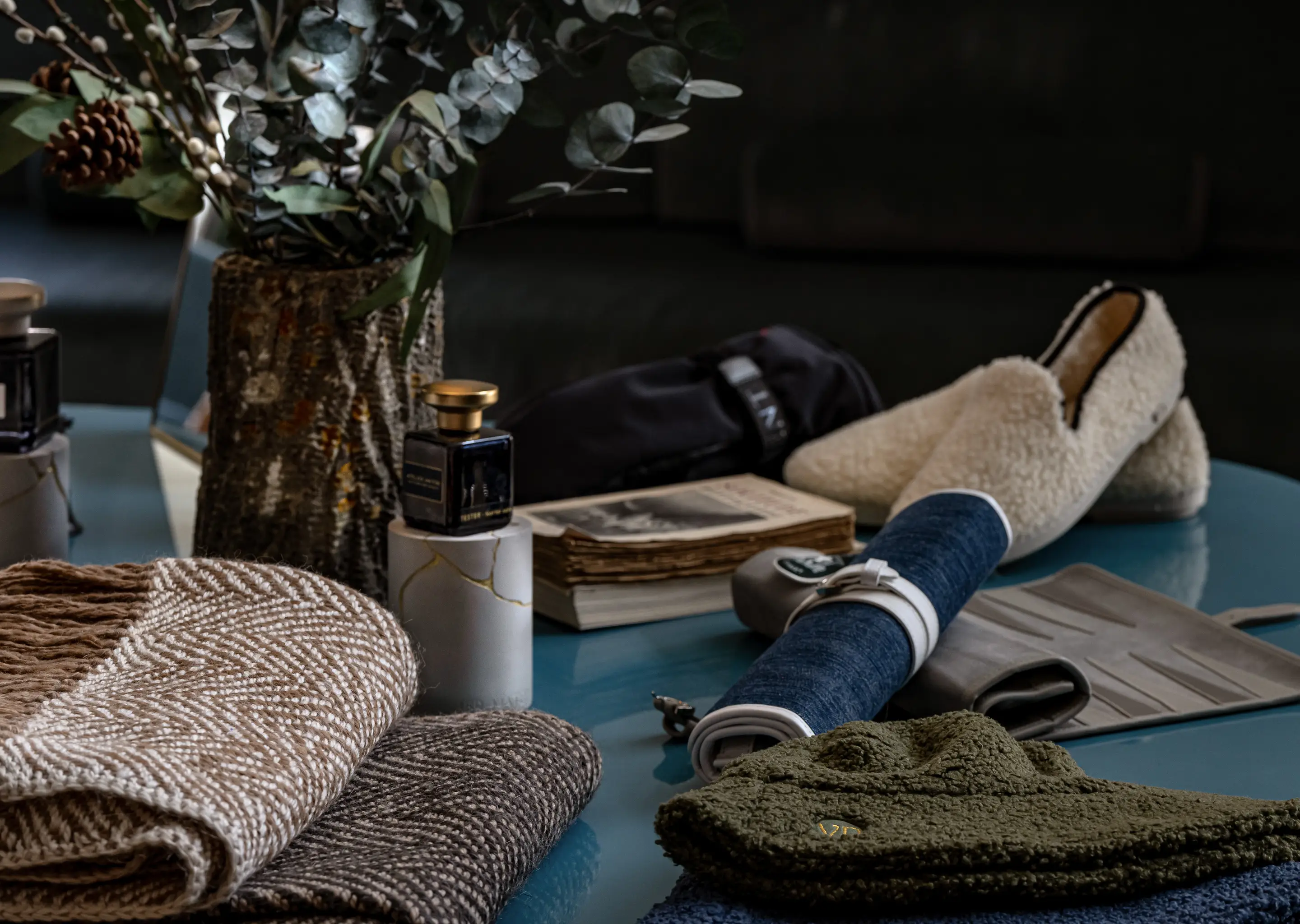 Arrangement of cozy wool scarves, a bottle of perfume, a dark-colored umbrella, an old book, beige slippers, and a green knit hat on a blue surface with eucalyptus branches in a textured vase.