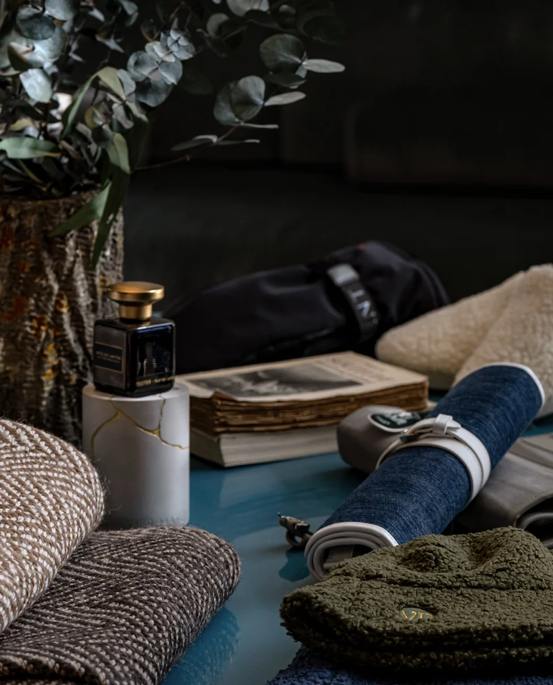 Assortment of textured fabrics, a small black perfume bottle on a white marble stand, a rolled-up piece of blue denim, an olive green knit hat, stacked vintage books, and greenery in a woven basket.