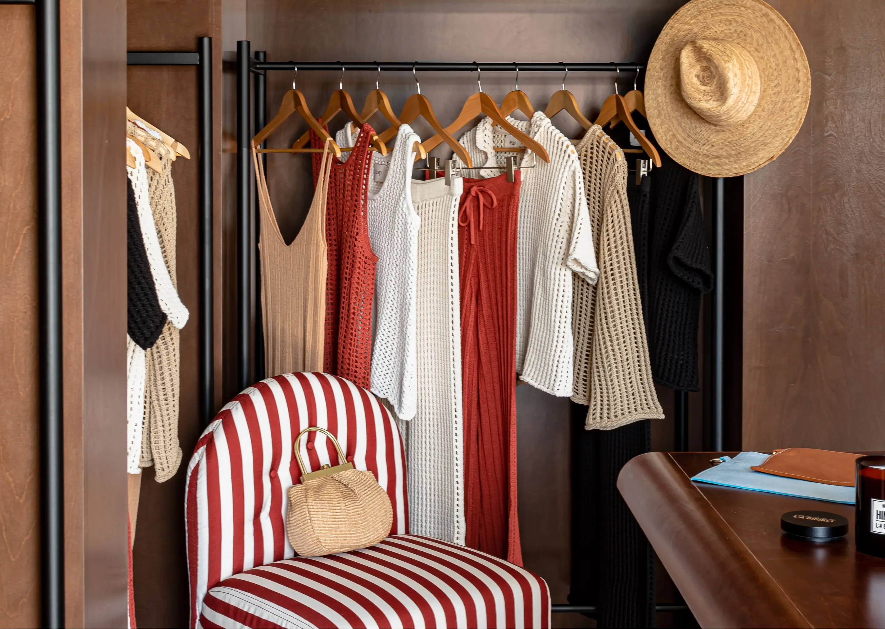 Clothing rack with knit dresses and tops in beige, red, white, and black, a straw hat hanging, and a red-and-white striped chair with a small woven handbag.