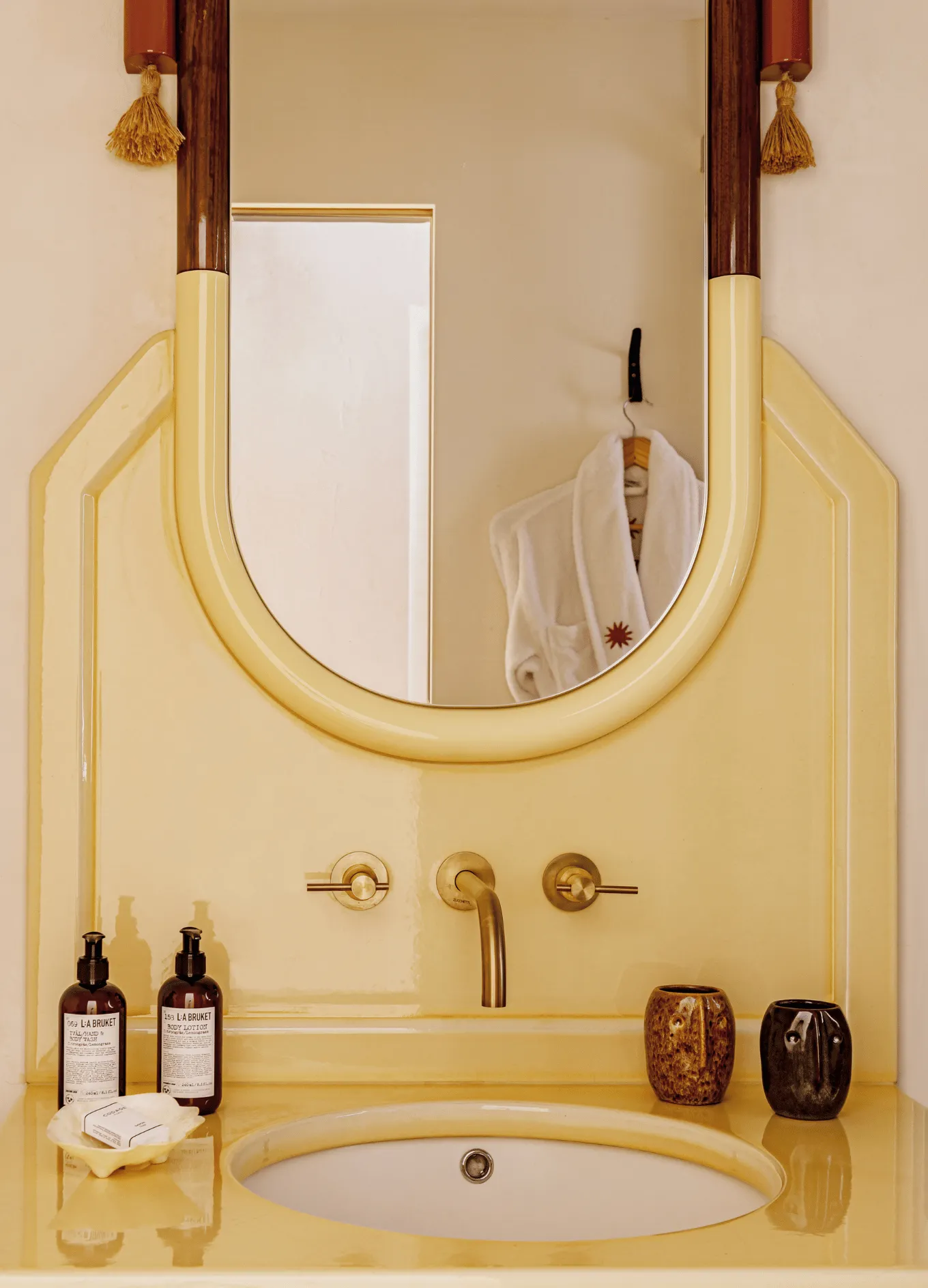 Yellow bathroom sink with gold faucet, oval mirror, lotion bottles, soap, and two artistic ceramic cups.