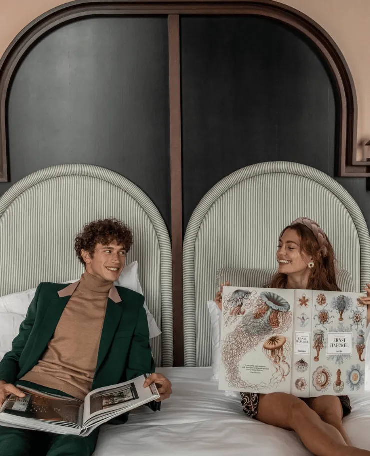 A man and a woman sitting on a bed with arched headboards, each holding an open book; the woman’s book features illustrations of jellyfish.