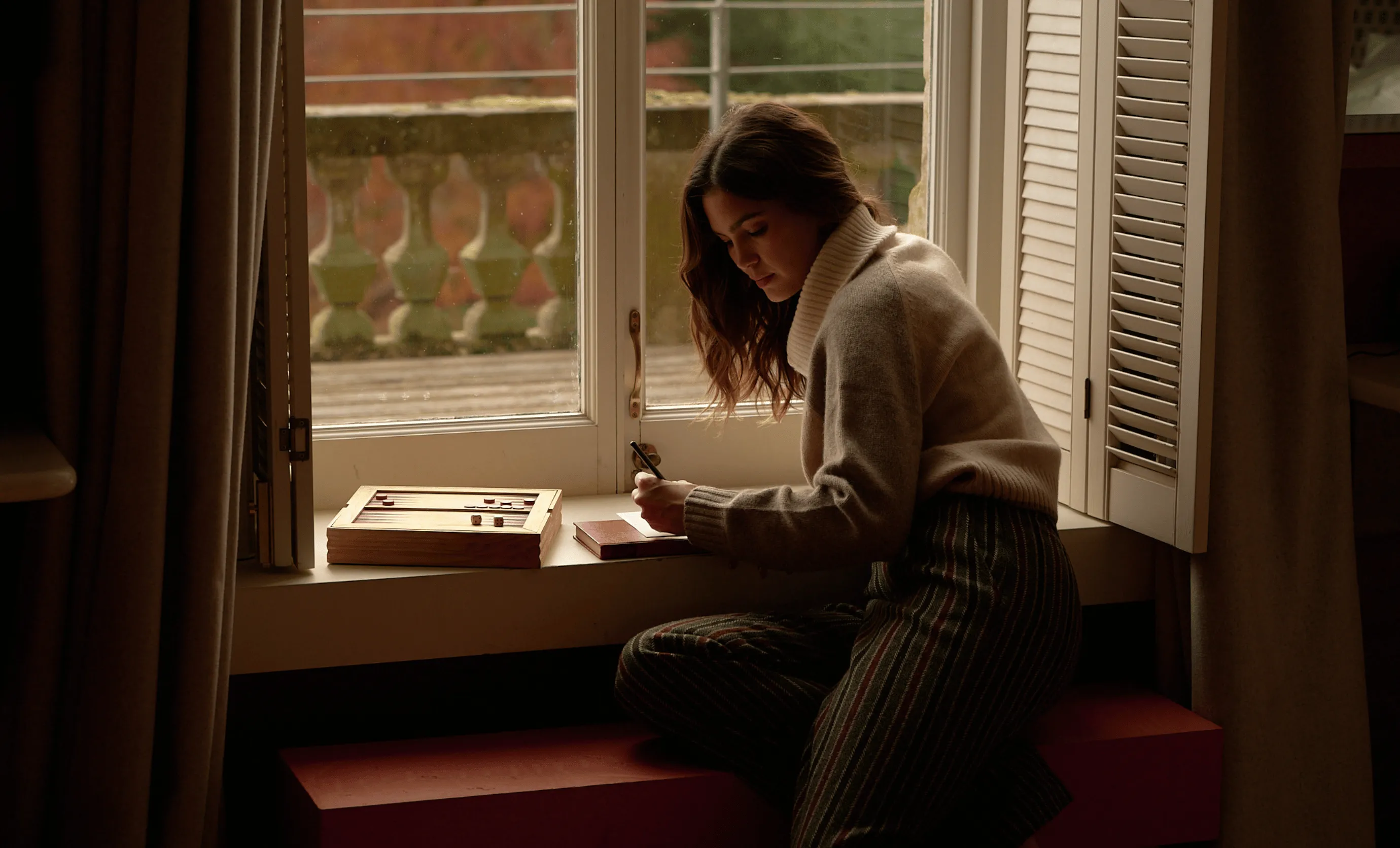 Woman in a beige turtleneck sweater sitting by a window, writing in a notebook with a wooden backgammon set beside her.