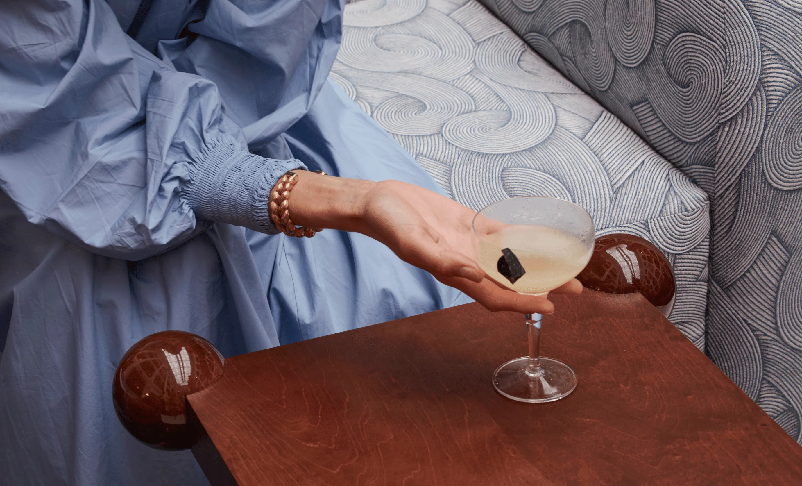 Hand with a gold bracelet holding a cocktail glass with a yellow drink and a small black garnish on a wooden table next to a patterned upholstered bench.