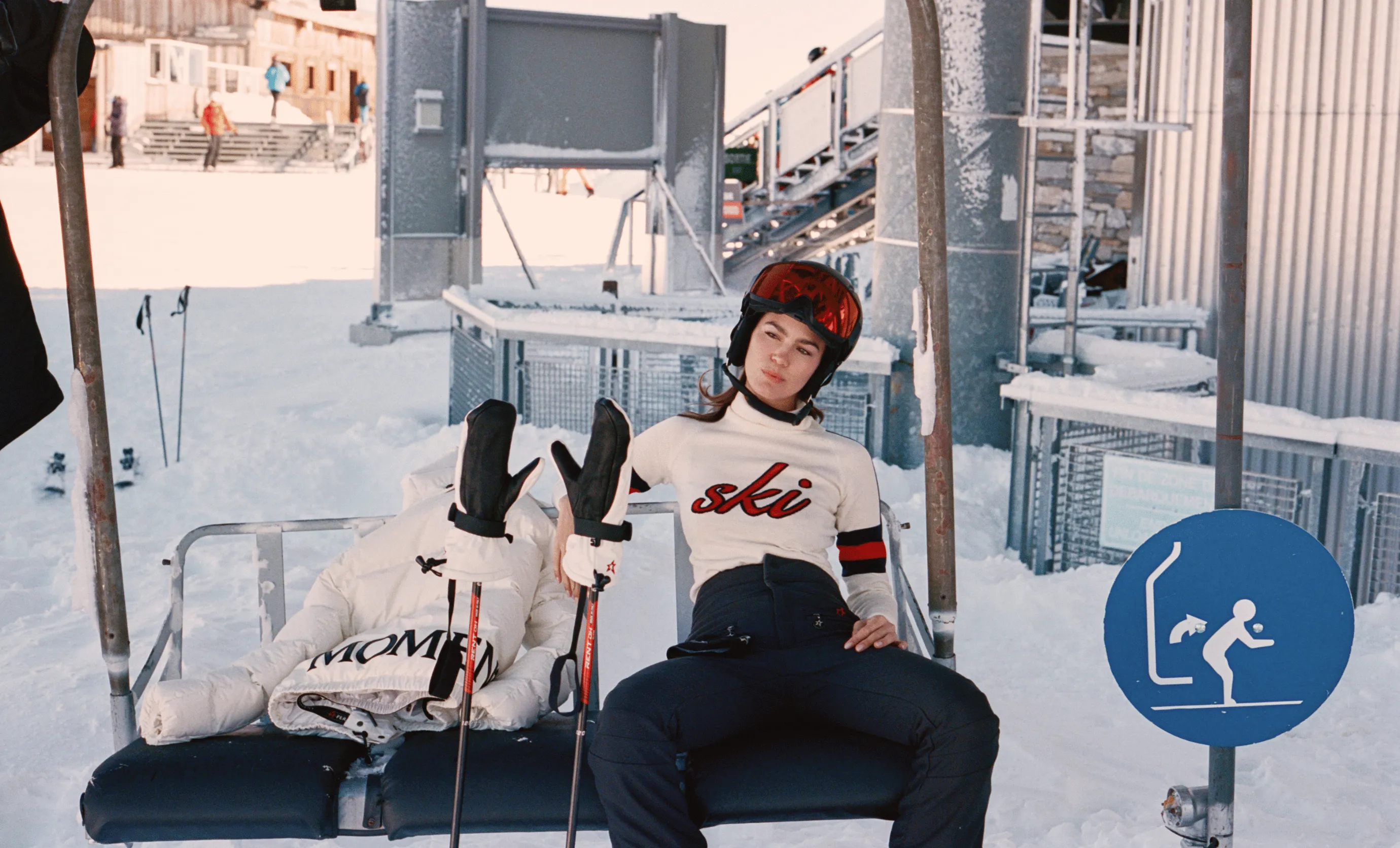Woman in white ski sweater and helmet sitting on ski lift chair next to white jacket and ski poles in snowy ski resort.
