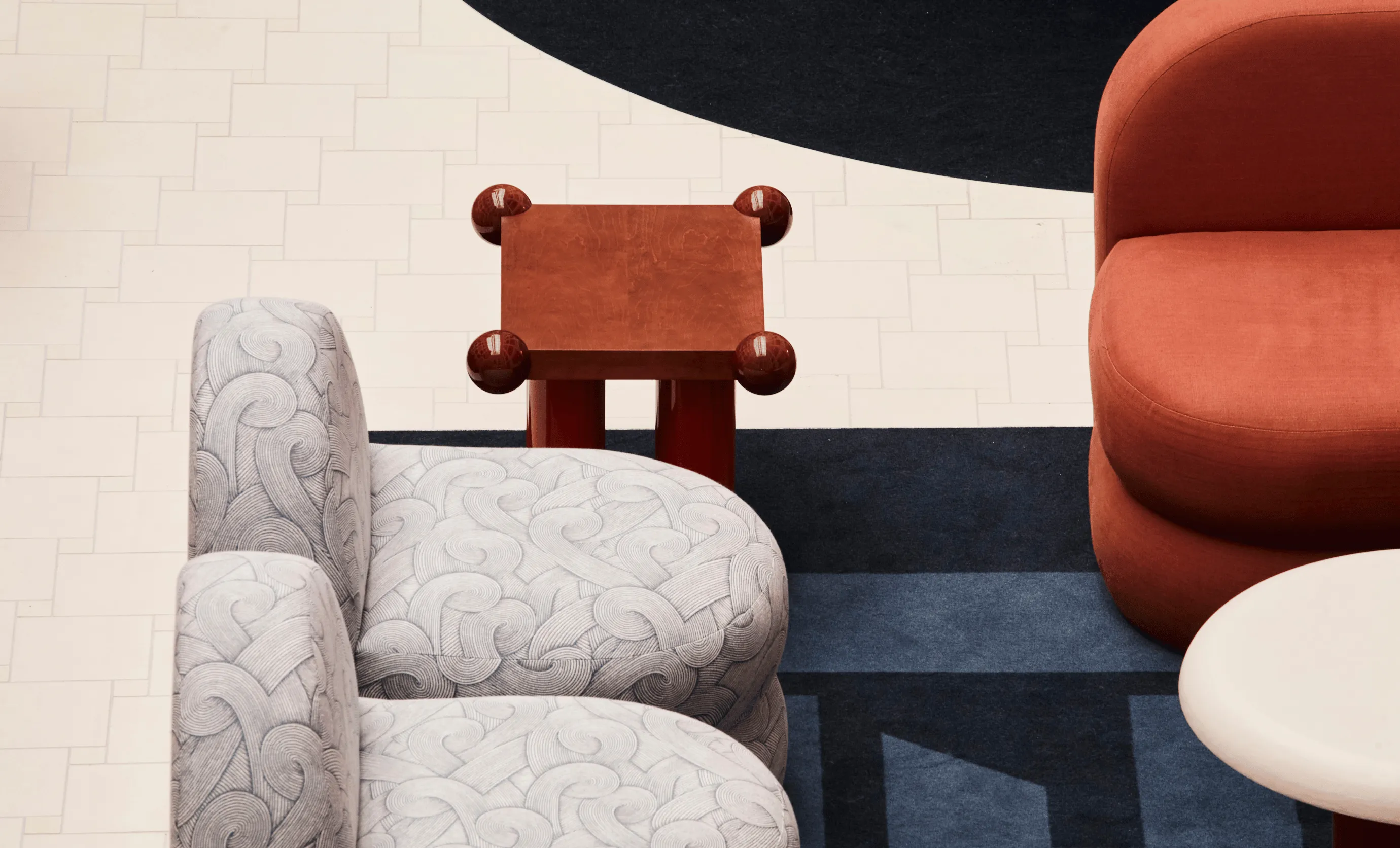 Modern lounge area with patterned light gray armchair, small red side table with spherical legs, rust-colored rounded sofa, and white circular table on a geometric dark blue rug.