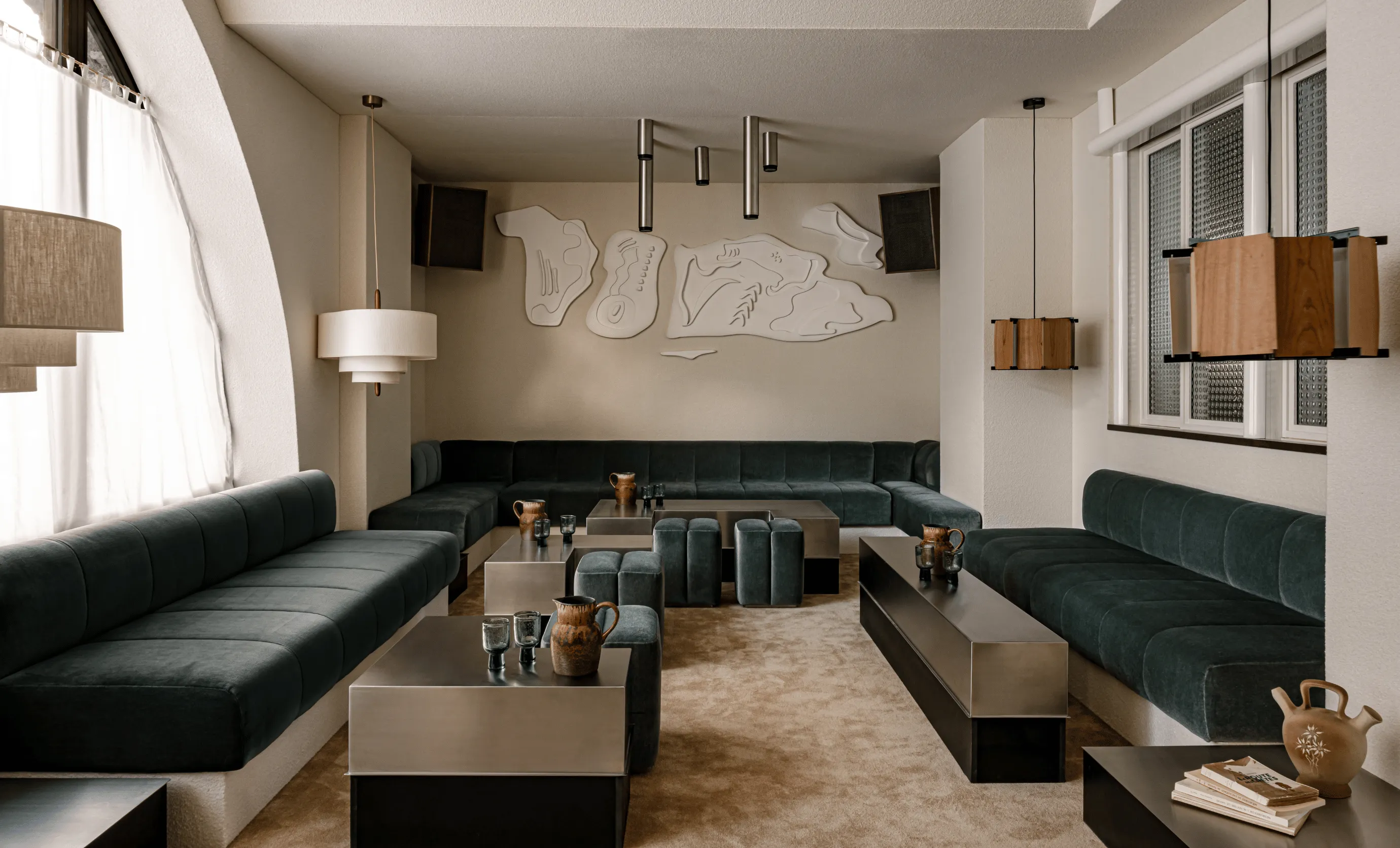 Modern hotel lounge with dark green velvet sofas arranged in a U shape around minimalist stainless steel tables, accented by ceramic pitchers and glassware, with abstract wall art and pendant lamps.