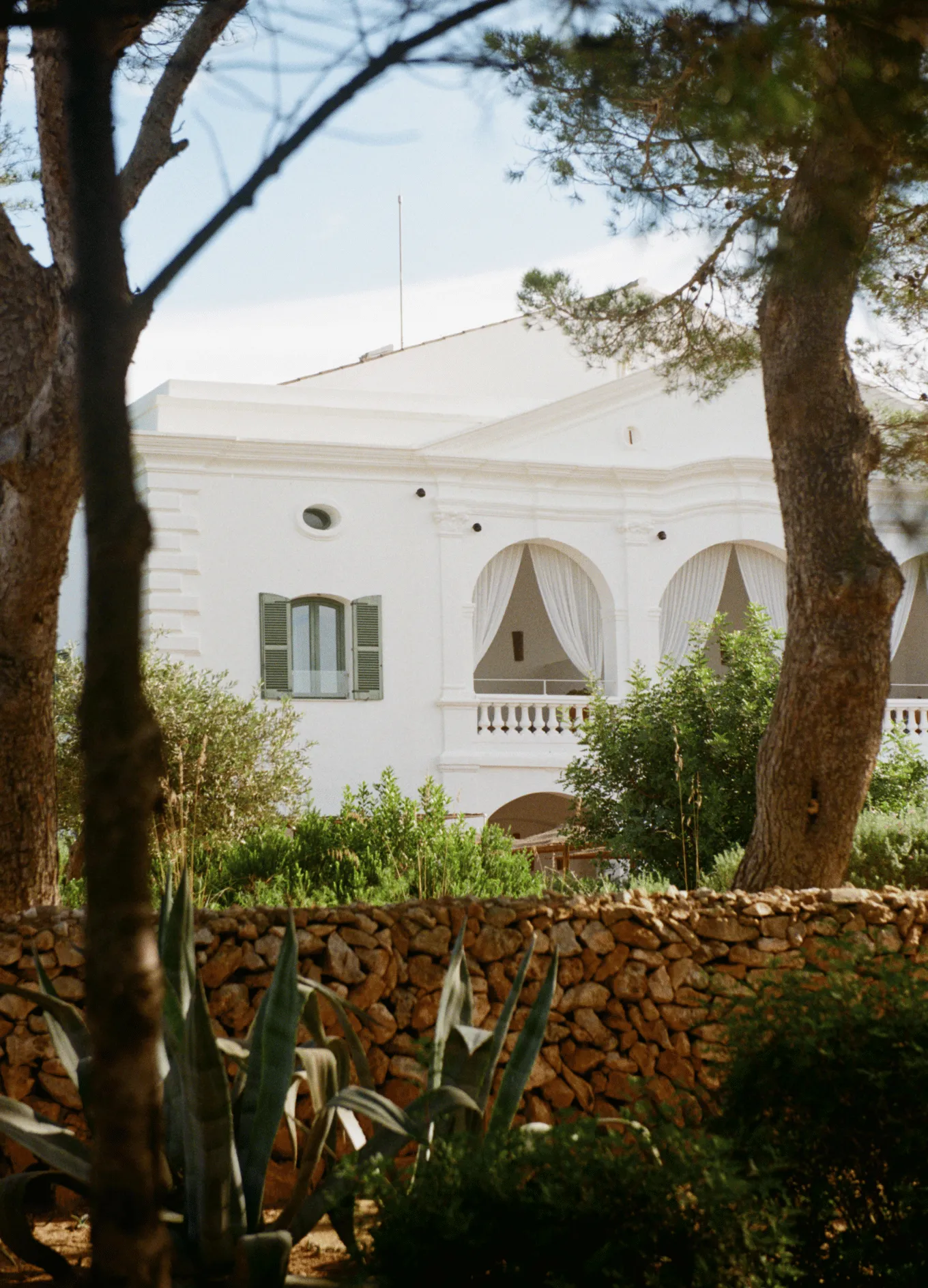 White building with green shuttered windows and arched balconies with curtains, partially obscured by trees and a stone wall in a garden setting.
