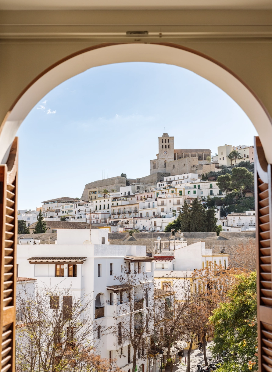 View of white buildings and a hill with a clock tower framed by an arched window with wooden shutters in Ibiza.