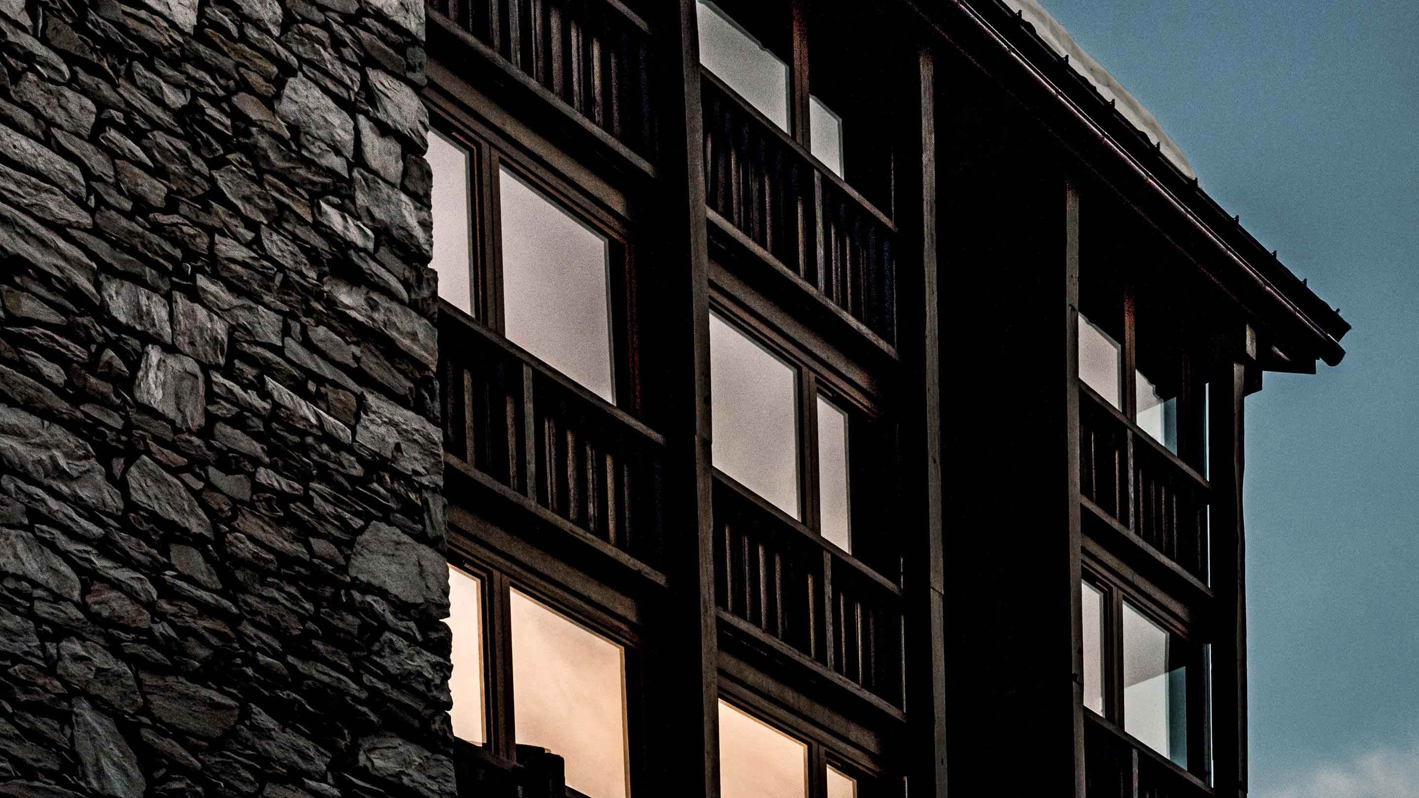 Close-up of a building corner with stone wall on the left and large dark-framed windows reflecting a cloudy sky.