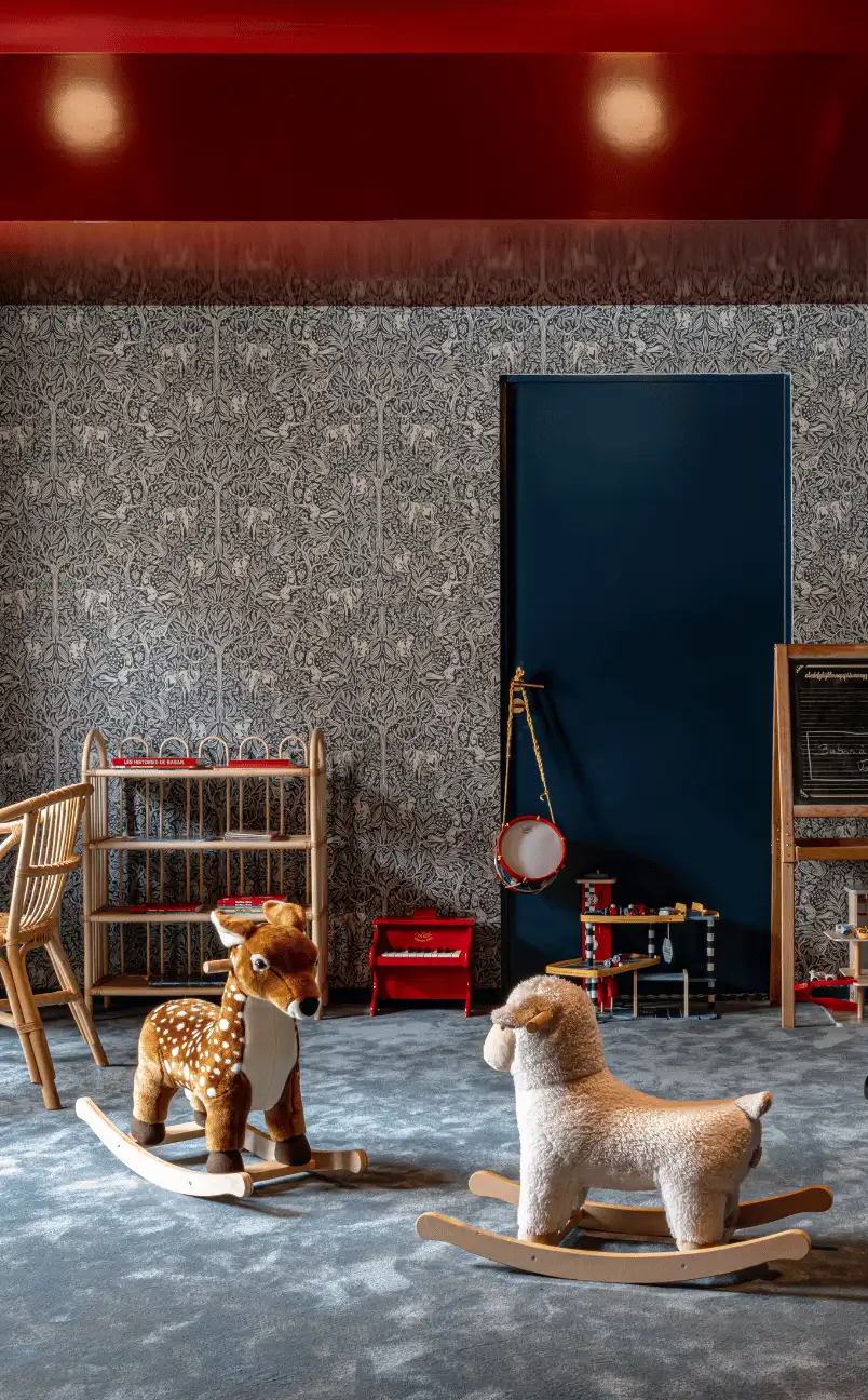 Children's playroom with a deer and a sheep rocking toy on a blue carpet, a wooden chair and shelves, a small red piano, and a blackboard against a patterned wallpaper and dark blue door.