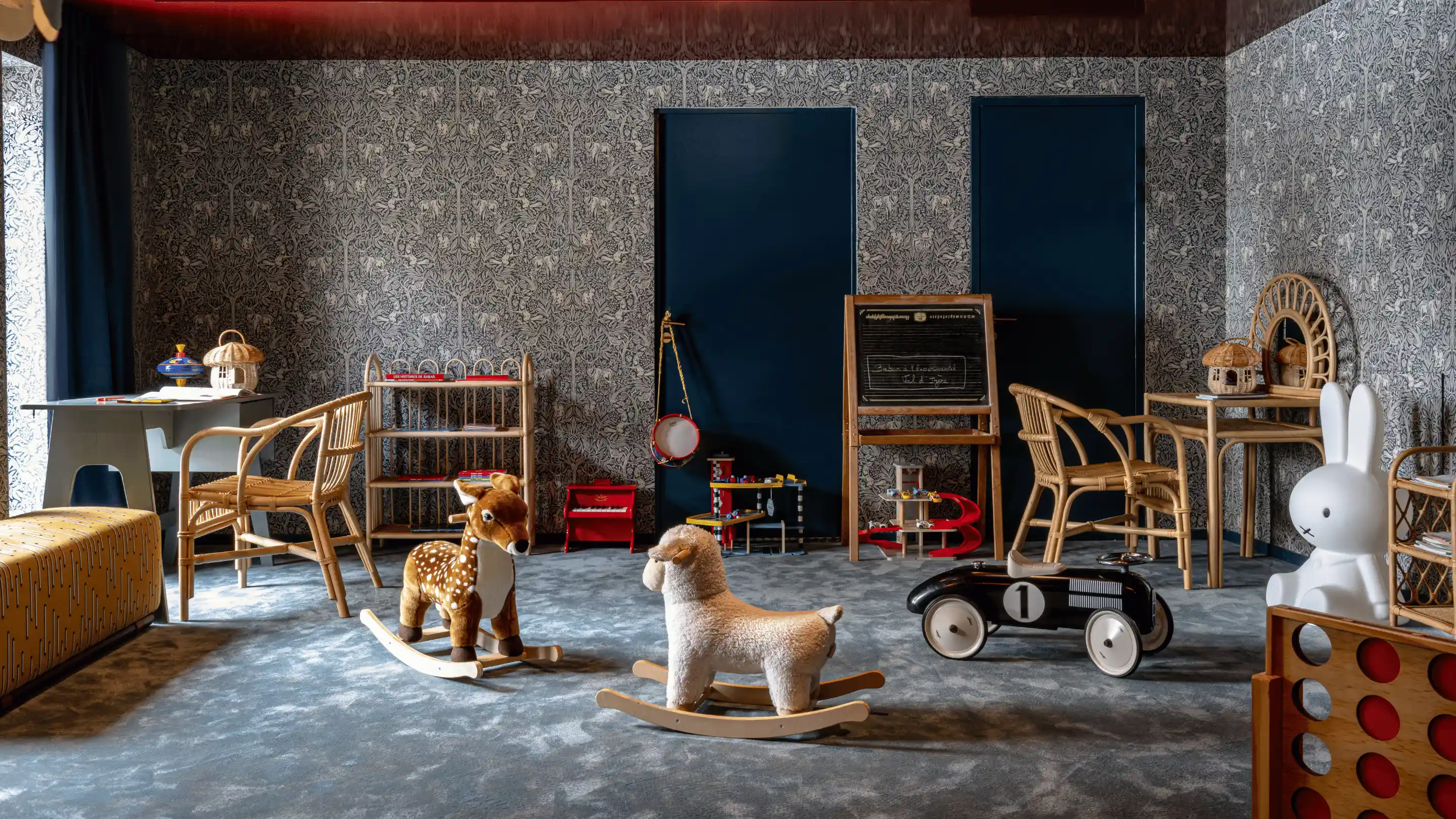 Children's playroom with two rocking animal toys, wicker furniture, toy car, chalkboard, and patterned wallpaper.