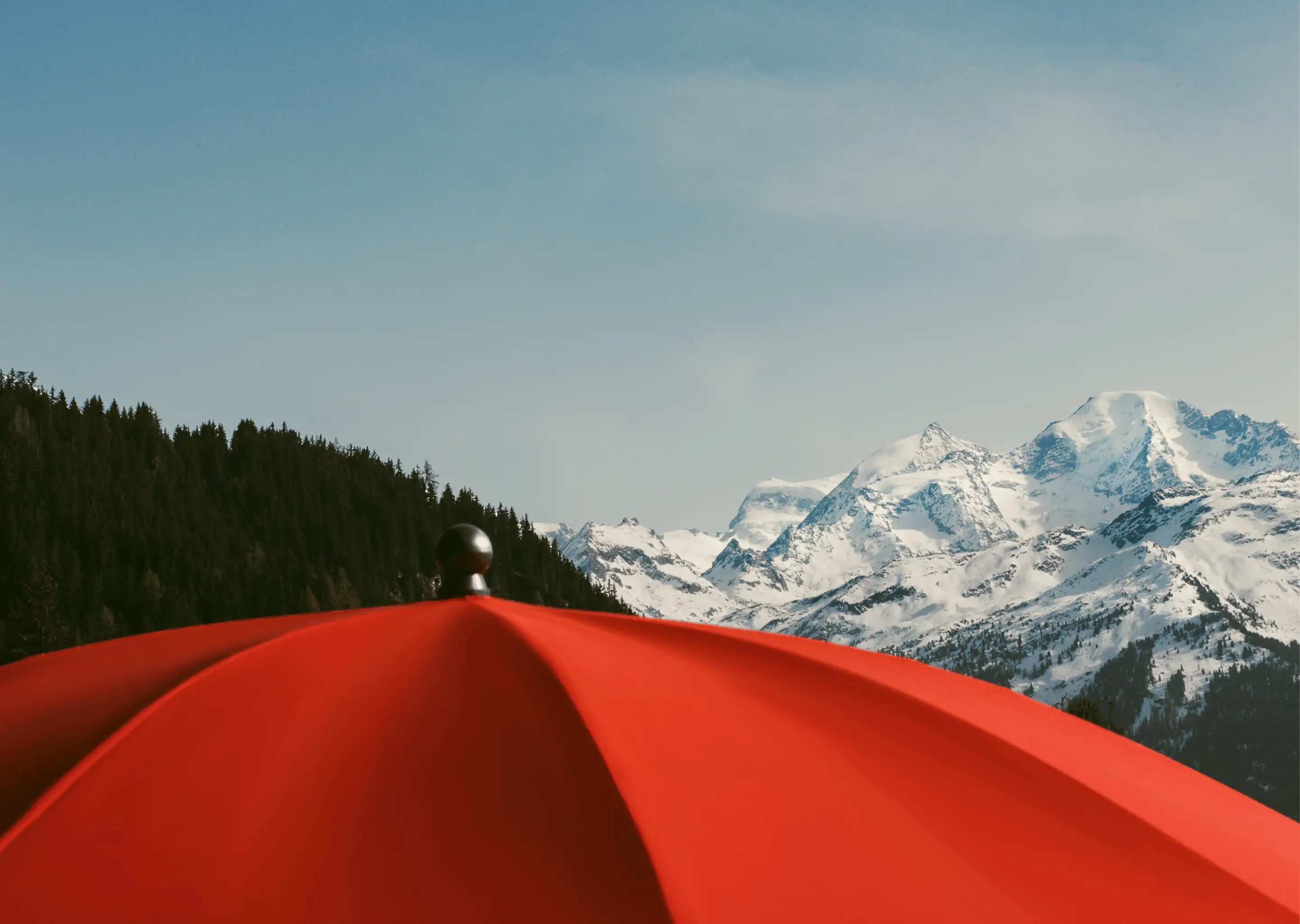 Red patio umbrella in the foreground with snow-covered mountains and pine trees under a blue sky in the background.