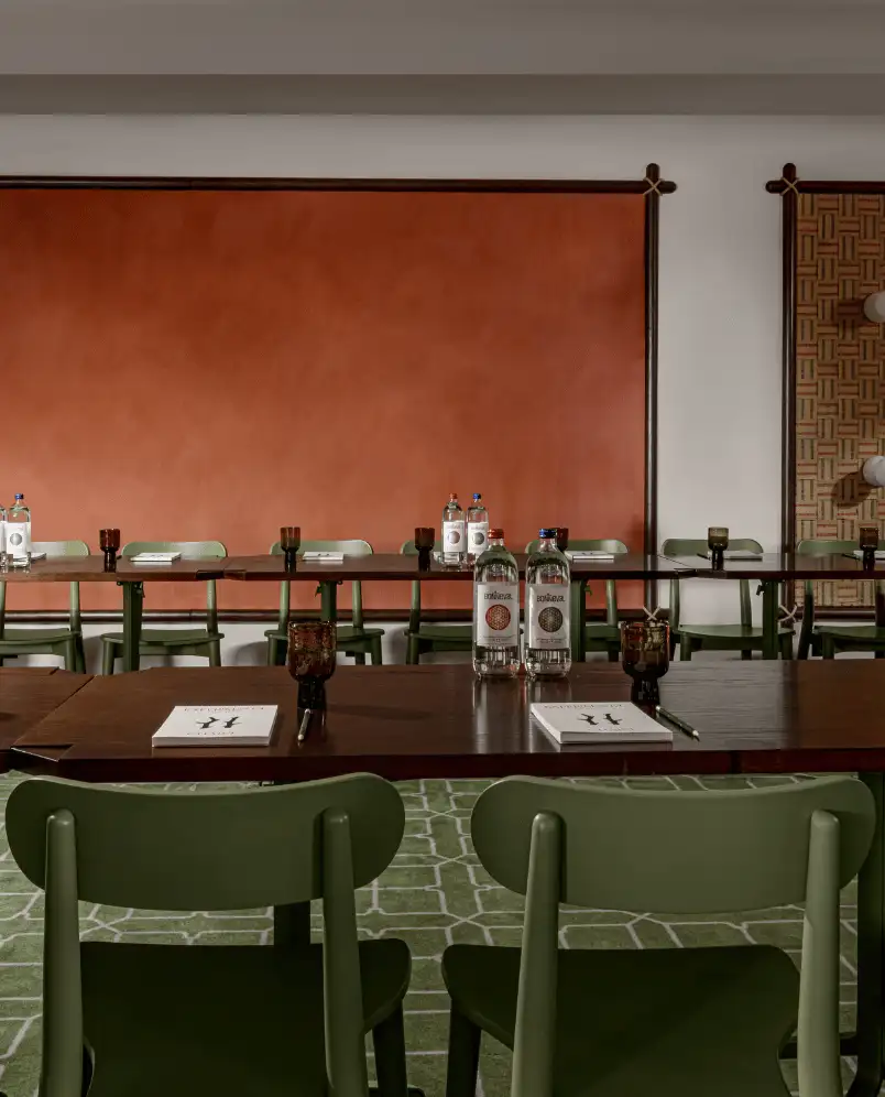 Conference room with green chairs around dark wooden tables, each set with notebooks, pens, and bottles of water against an orange and patterned wall backdrop.