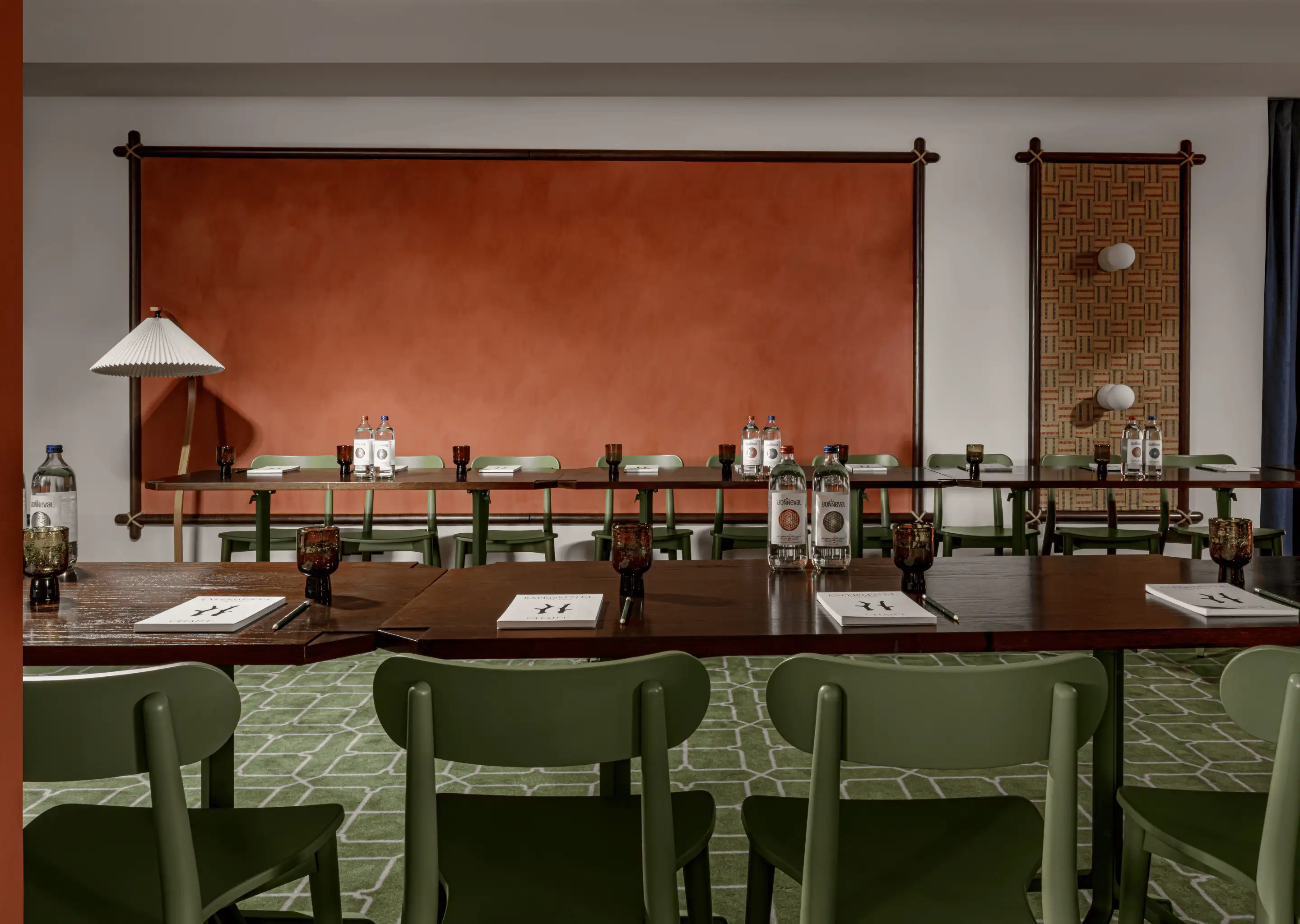 Neat meeting room with wooden tables, green chairs, bottles of water, notebooks, and pens against an orange textured wall and patterned carpet.