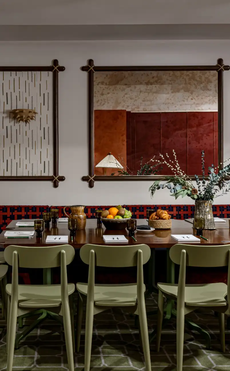 Dining table set with green chairs, amber glassware, bowls of fruit and croissants, notebooks, and a vase with branches and greenery in a stylish room with framed mirror and wall art.