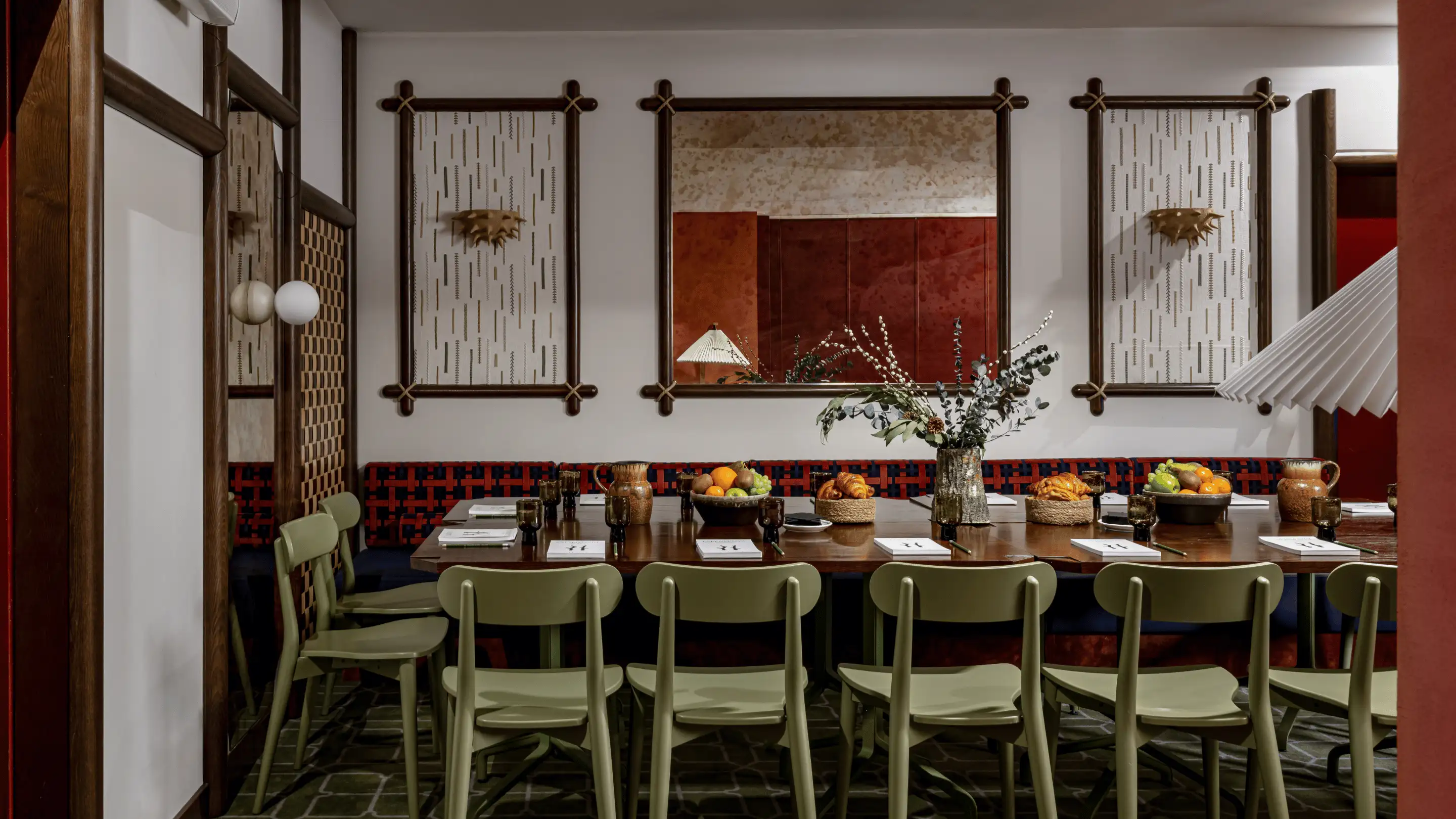 Dining room table set with green chairs, decorated with fruit bowls, croissants, and flower arrangements under framed wall art and a large mirror.