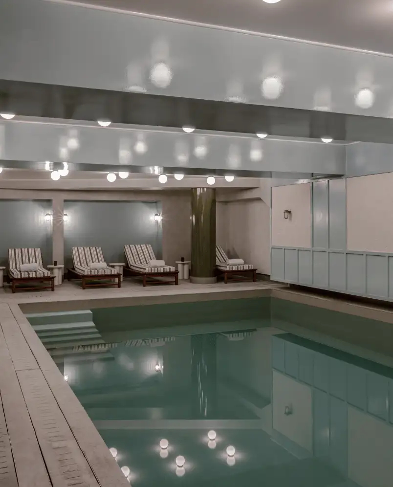 Indoor swimming pool with four striped lounge chairs and soft overhead lighting reflecting on the water.