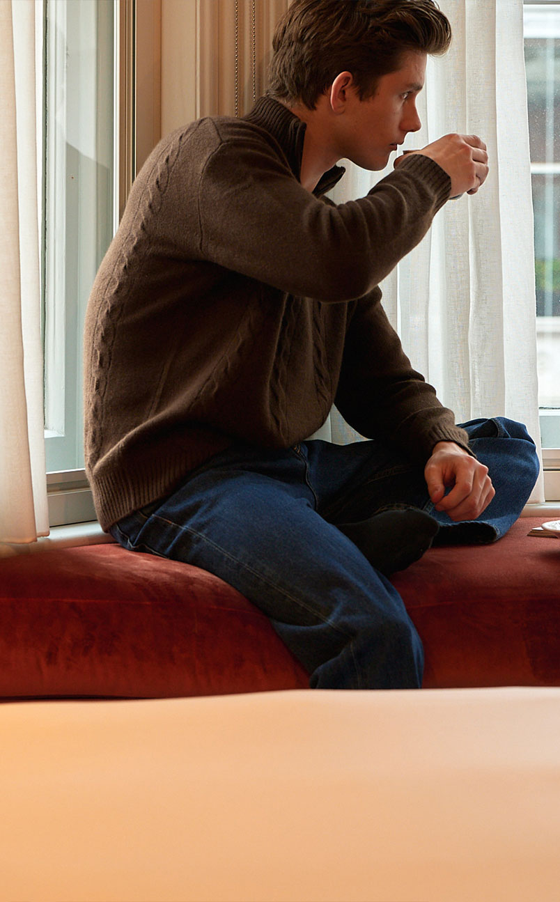 Young man sitting cross-legged on a window seat drinking from a small cup, looking outside through white curtains.