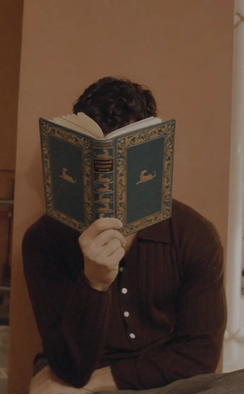 Person with curly hair wearing a dark brown shirt reads a green hardcover book with gold detailing, covering their face.