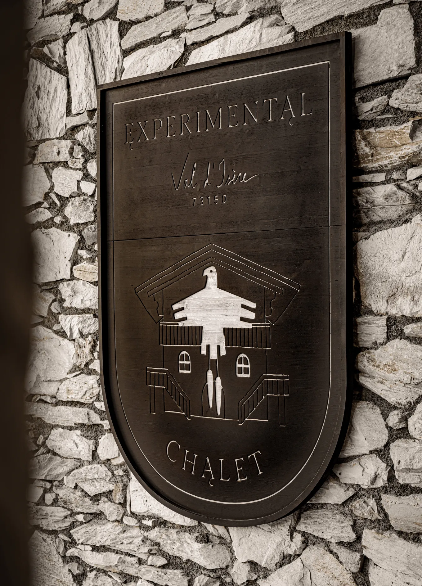 Wooden plaque mounted on a stone wall with the text Experimental Chalet and an engraved design of a stylized bird over a house sketch.