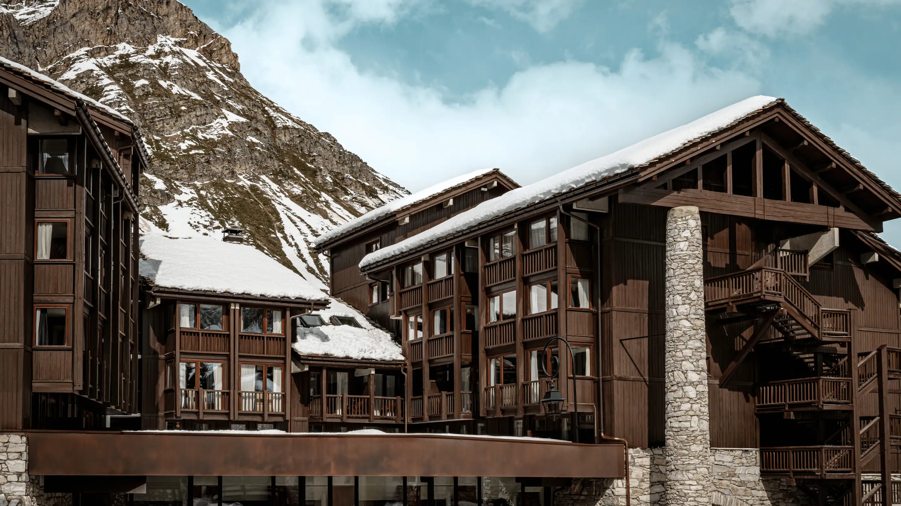 Snow-covered wooden chalet buildings in front of a rocky mountain under a partly cloudy sky.