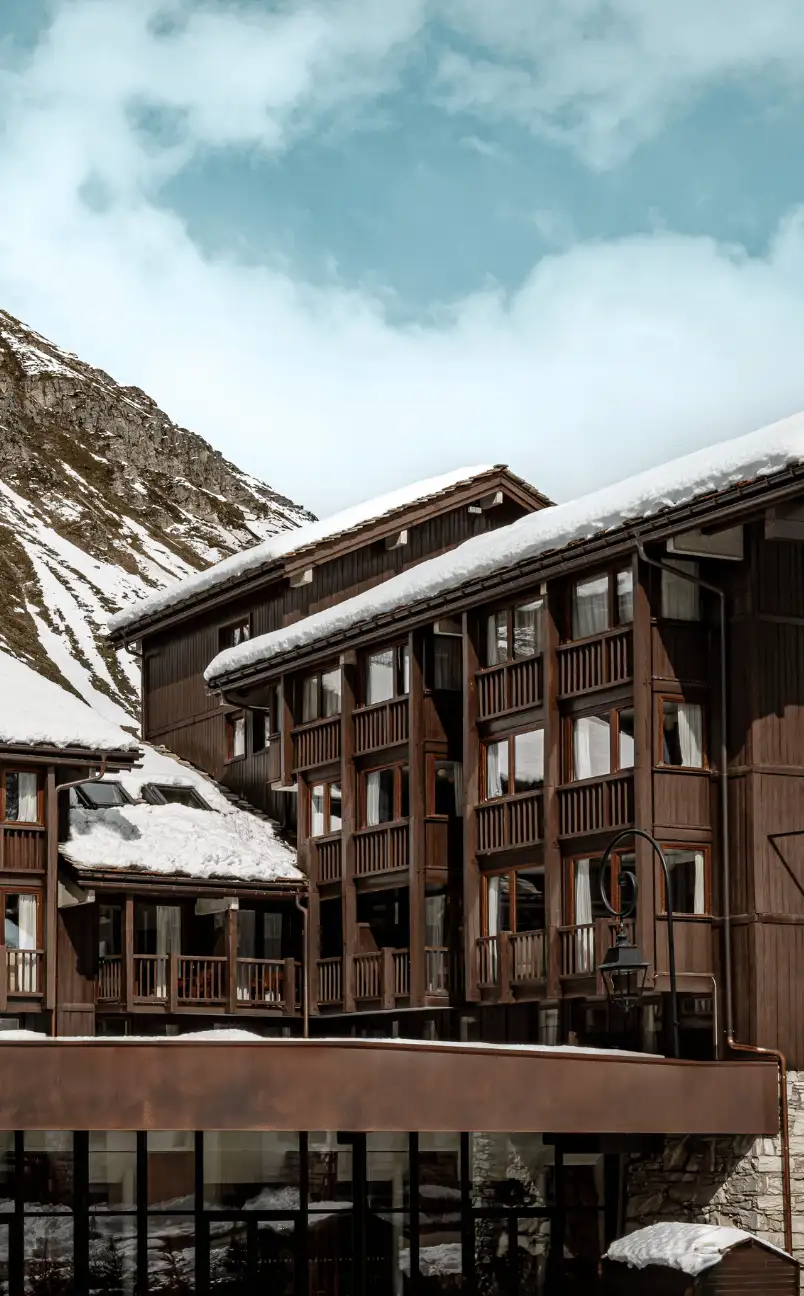 Snow-covered wooden alpine hotel buildings with balconies under a partly cloudy blue sky and mountain slope.