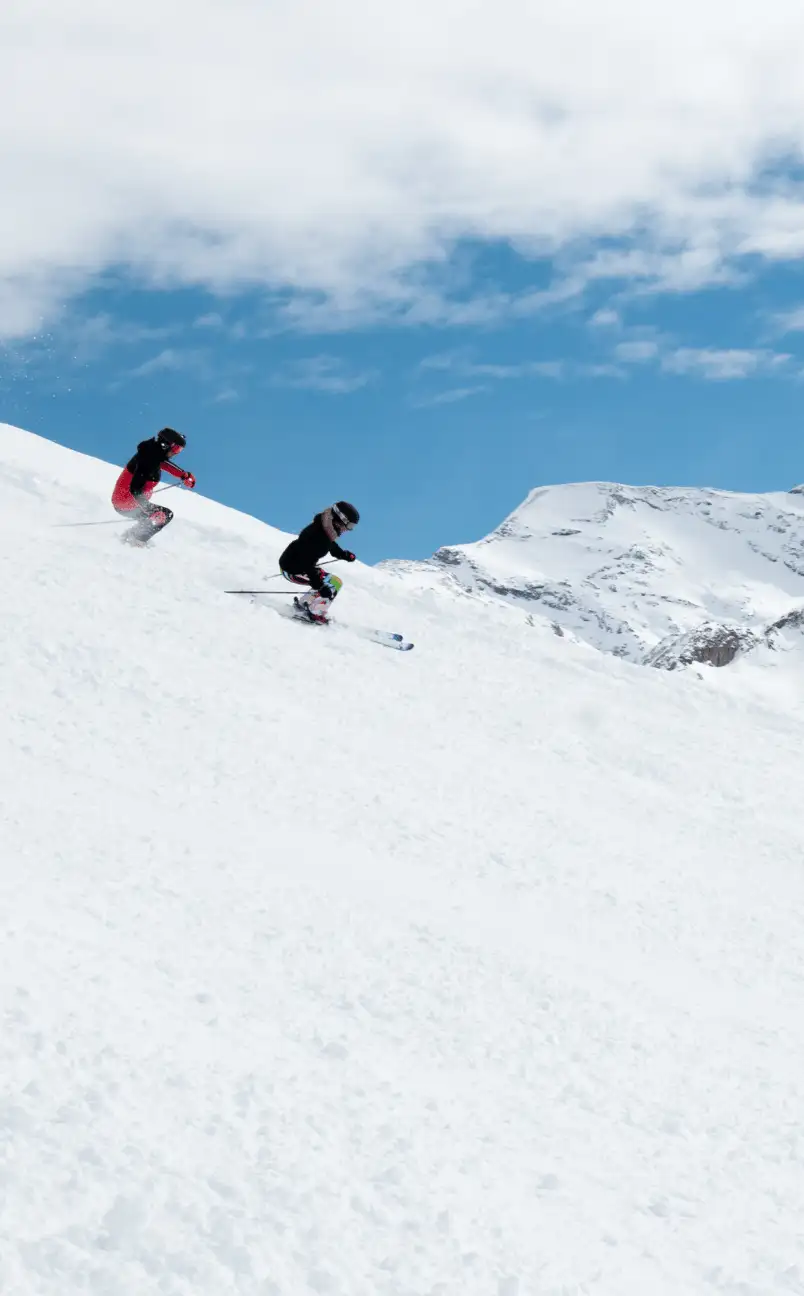 Two skiers wearing helmets and winter gear skiing down a snowy mountain slope under a partly cloudy blue sky.