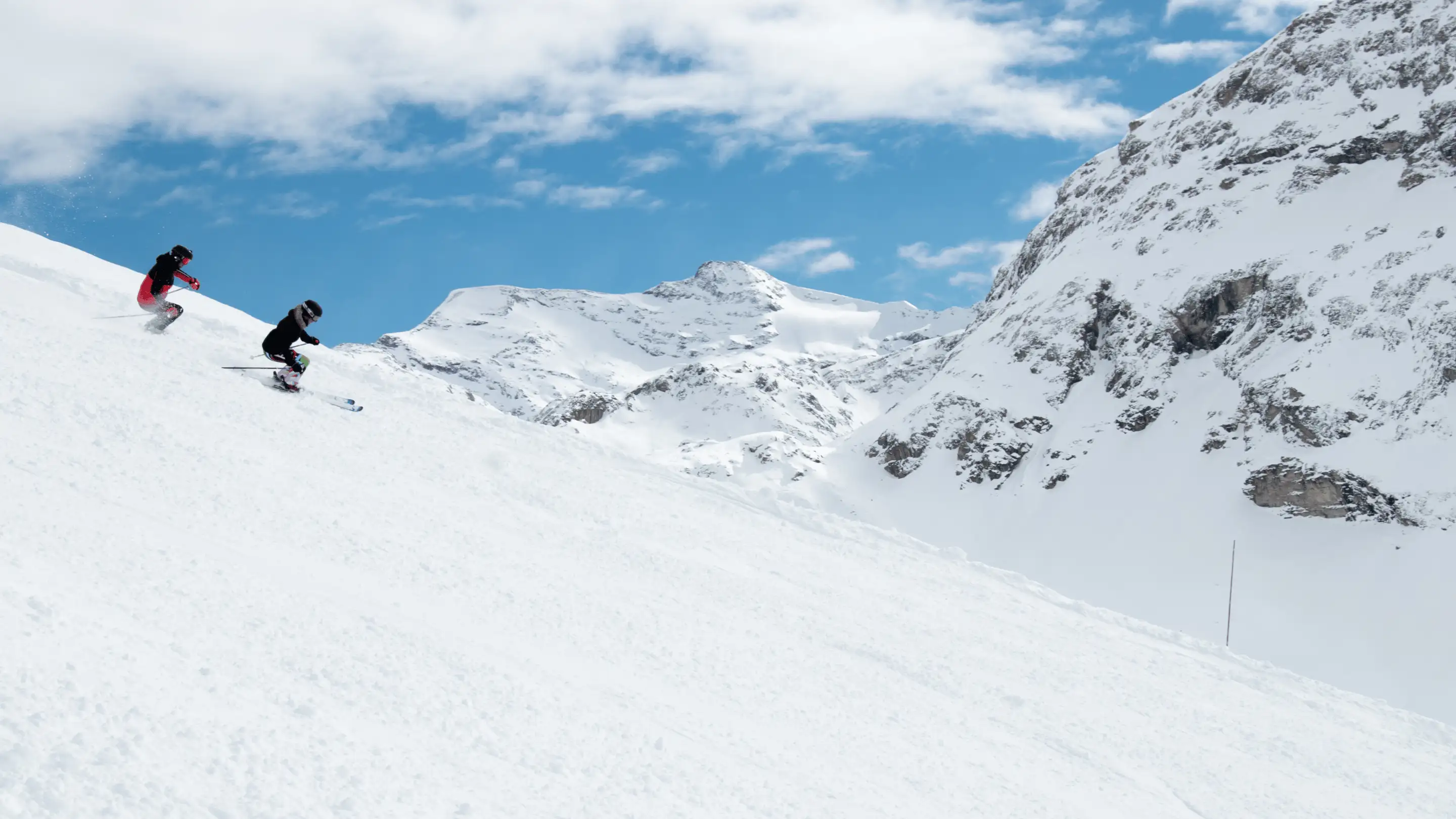 Two skiers descending a snowy slope with snow-covered mountains and blue sky in the background.