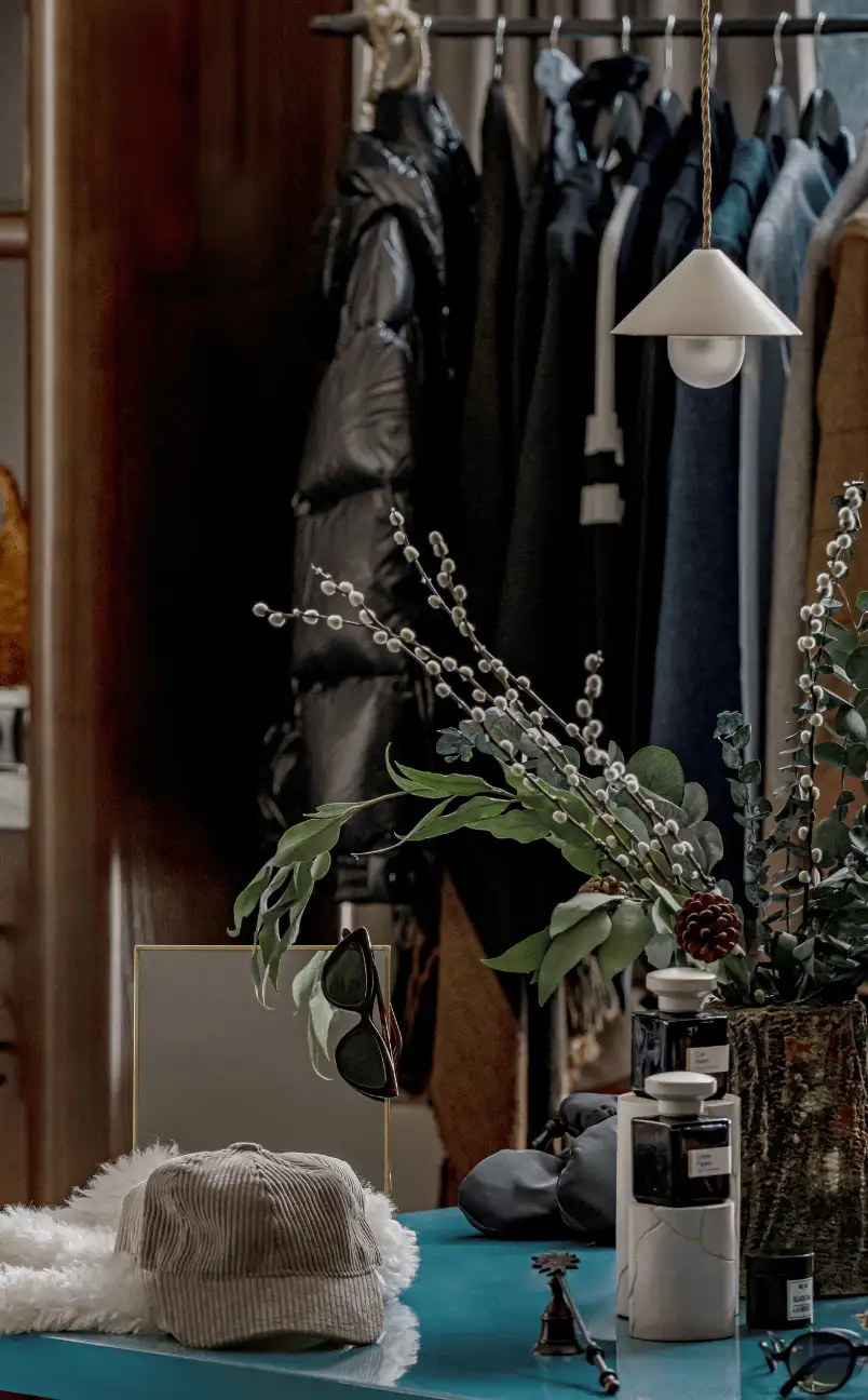 Table with corduroy cap, sunglasses, two perfume bottles, and decorative foliage vase, with winter coats on hangers in the background.