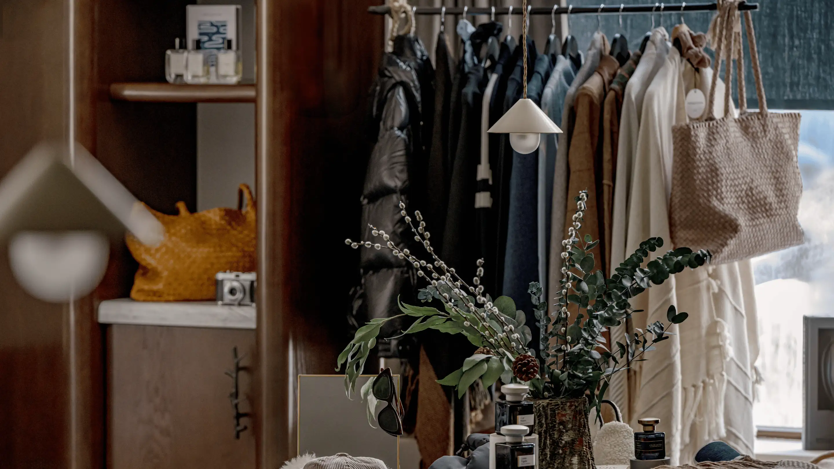 Cozy boutique display with hanging jackets, a woven bag, candles, sunglasses, and a leafy floral arrangement.