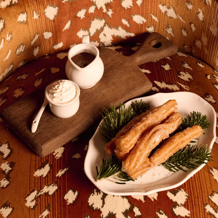 Plate of sugared churros garnished with green pine sprigs, served with whipped cream and chocolate sauce on a wooden board.