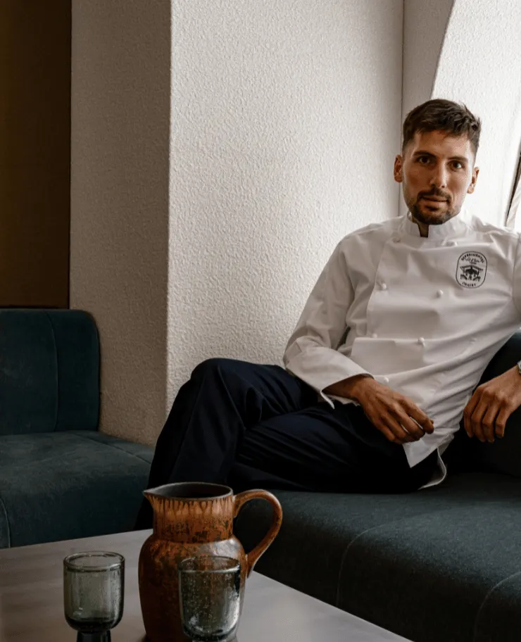Man in white chef jacket sitting on a dark green sofa beside a table with a brown pitcher and two glasses.