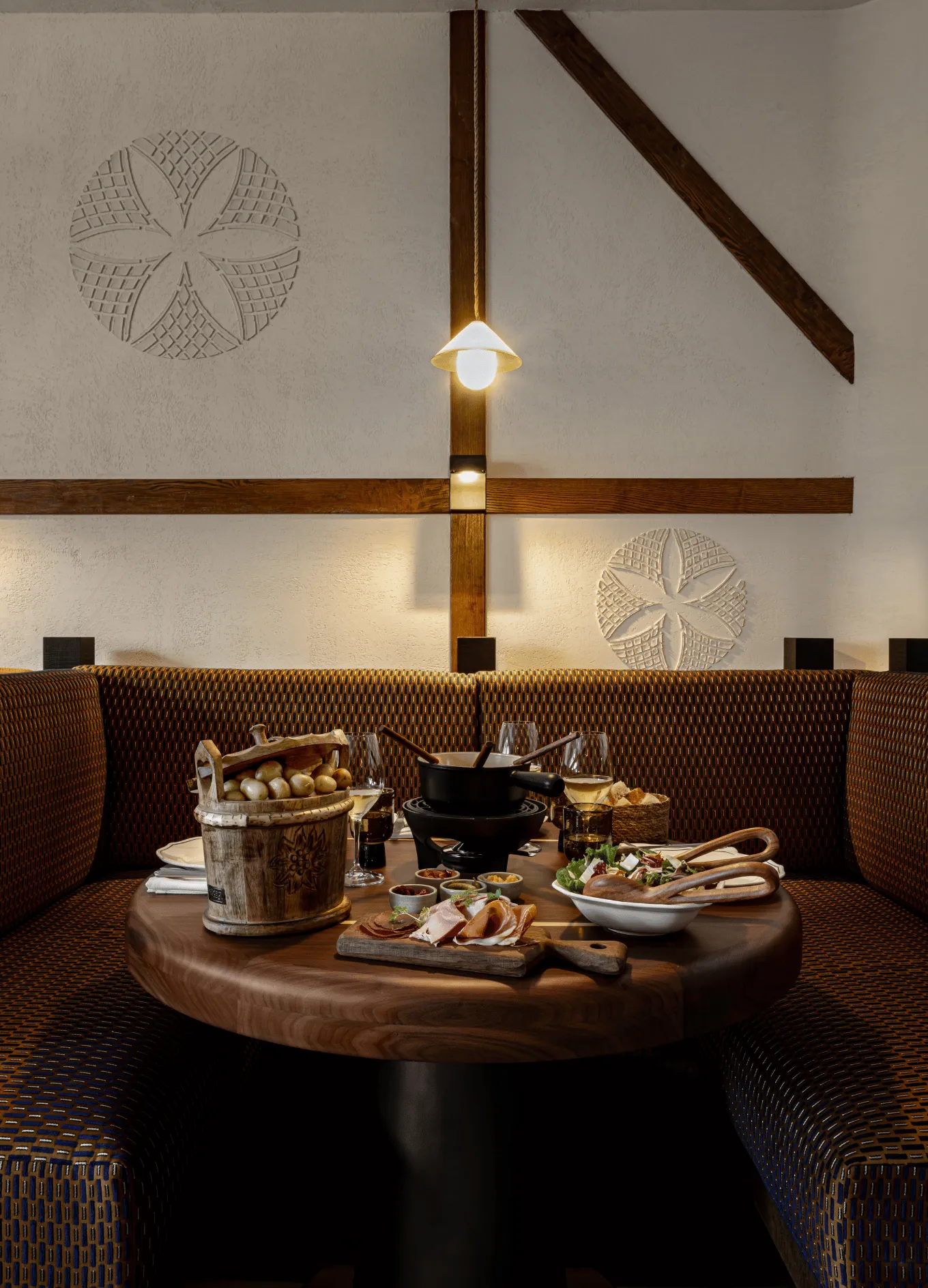 Cozy restaurant booth with patterned upholstery, wooden table set with fondue pot, sliced meats, salad, bread basket, and wine glasses under warm wall lighting.