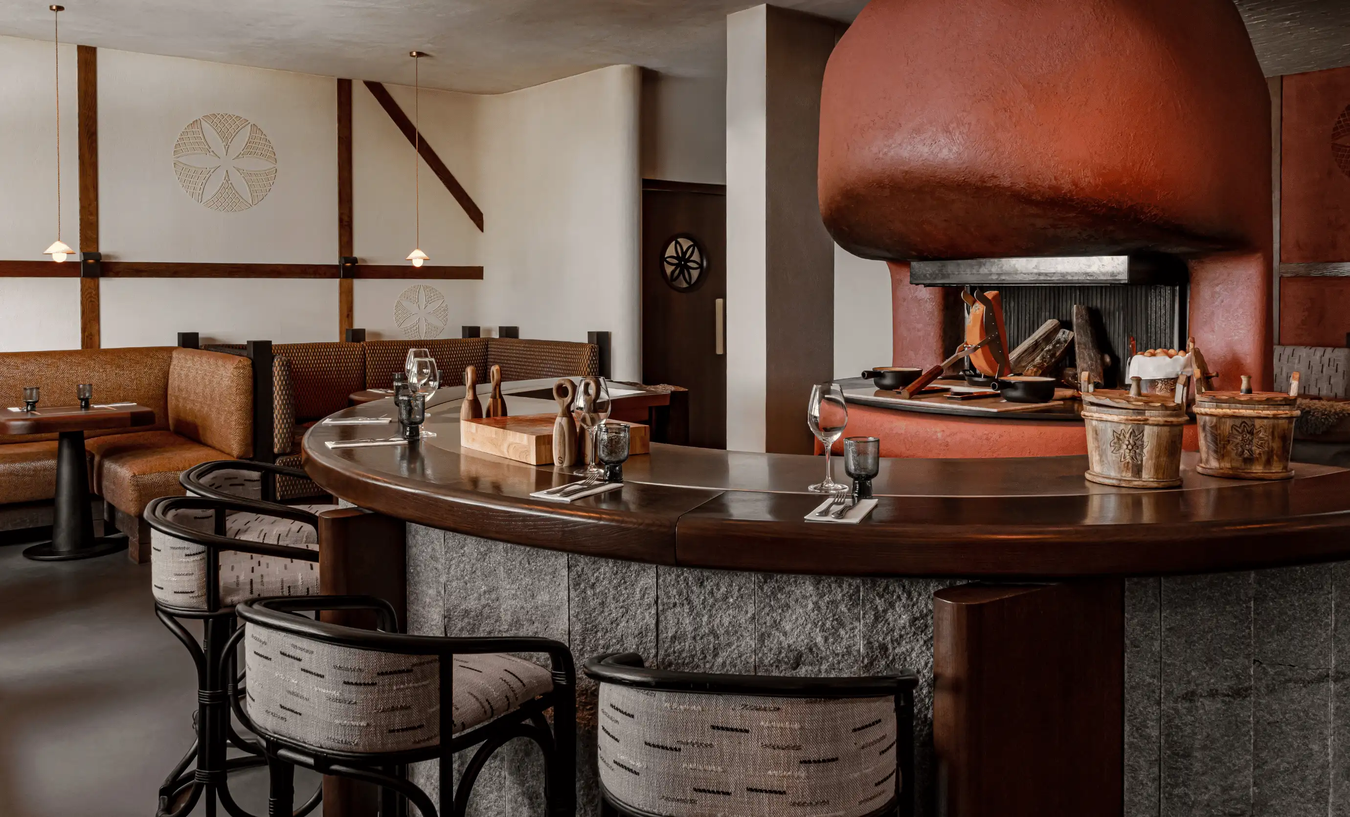 Cozy restaurant interior with curved wooden bar, upholstered chairs, and a large red fireplace with cooking utensils.