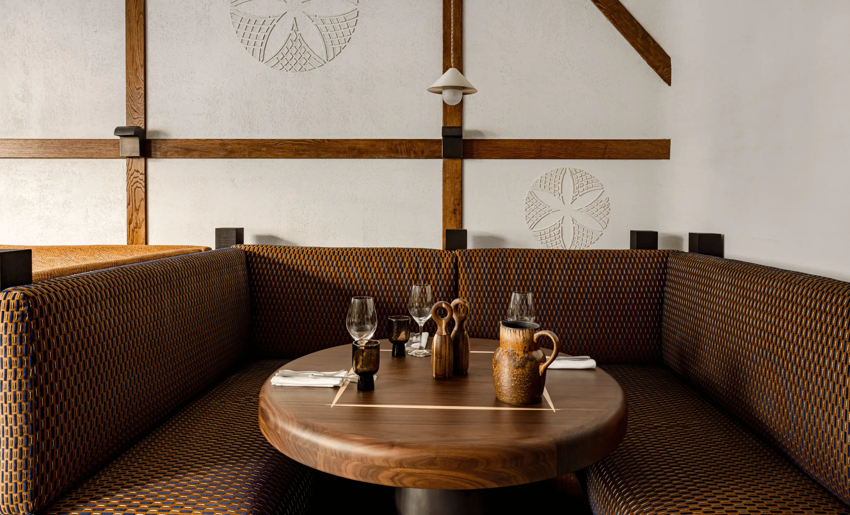 Cozy corner seating with patterned upholstery around a round wooden table set with glassware, napkins, wooden salt and pepper shakers, and a rustic ceramic pitcher.