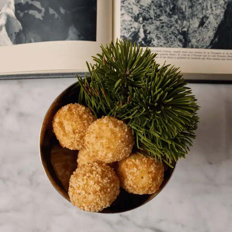 Top view of a bowl containing round, crumb-coated snacks and a bunch of green pine needles, with an open book in the background.
