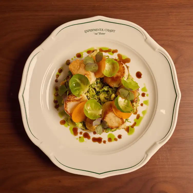 Plate with seared scallops, Brussels sprout leaves, orange vegetable slices, and green and brown sauce drizzles, served on a white plate labeled Experimental Chalet Val d'Isère.