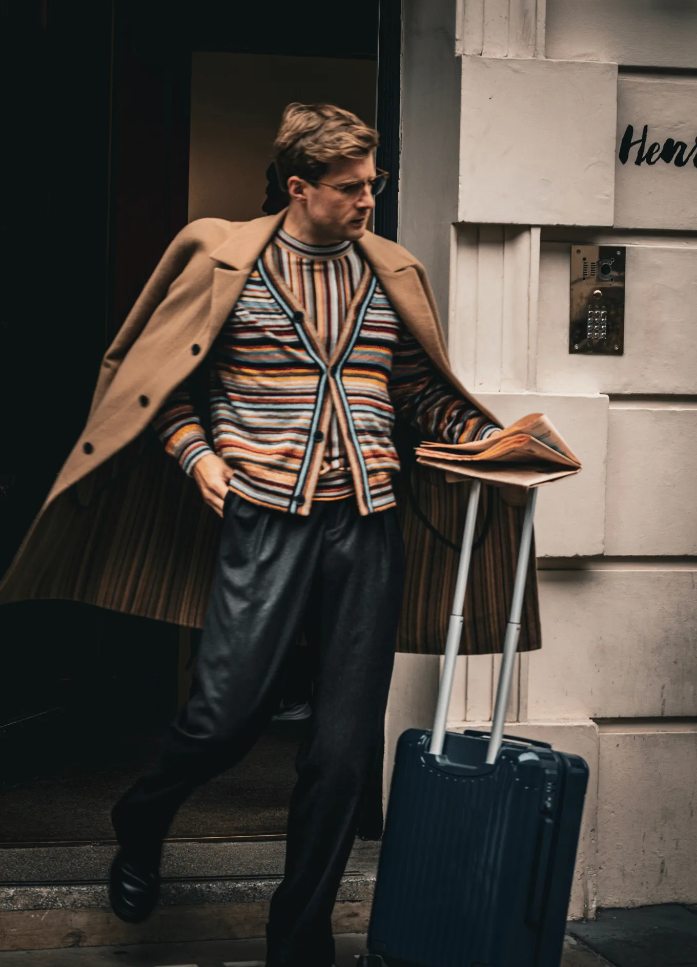Man wearing a striped cardigan and camel coat, pulling a navy blue rolling suitcase and holding folded papers.