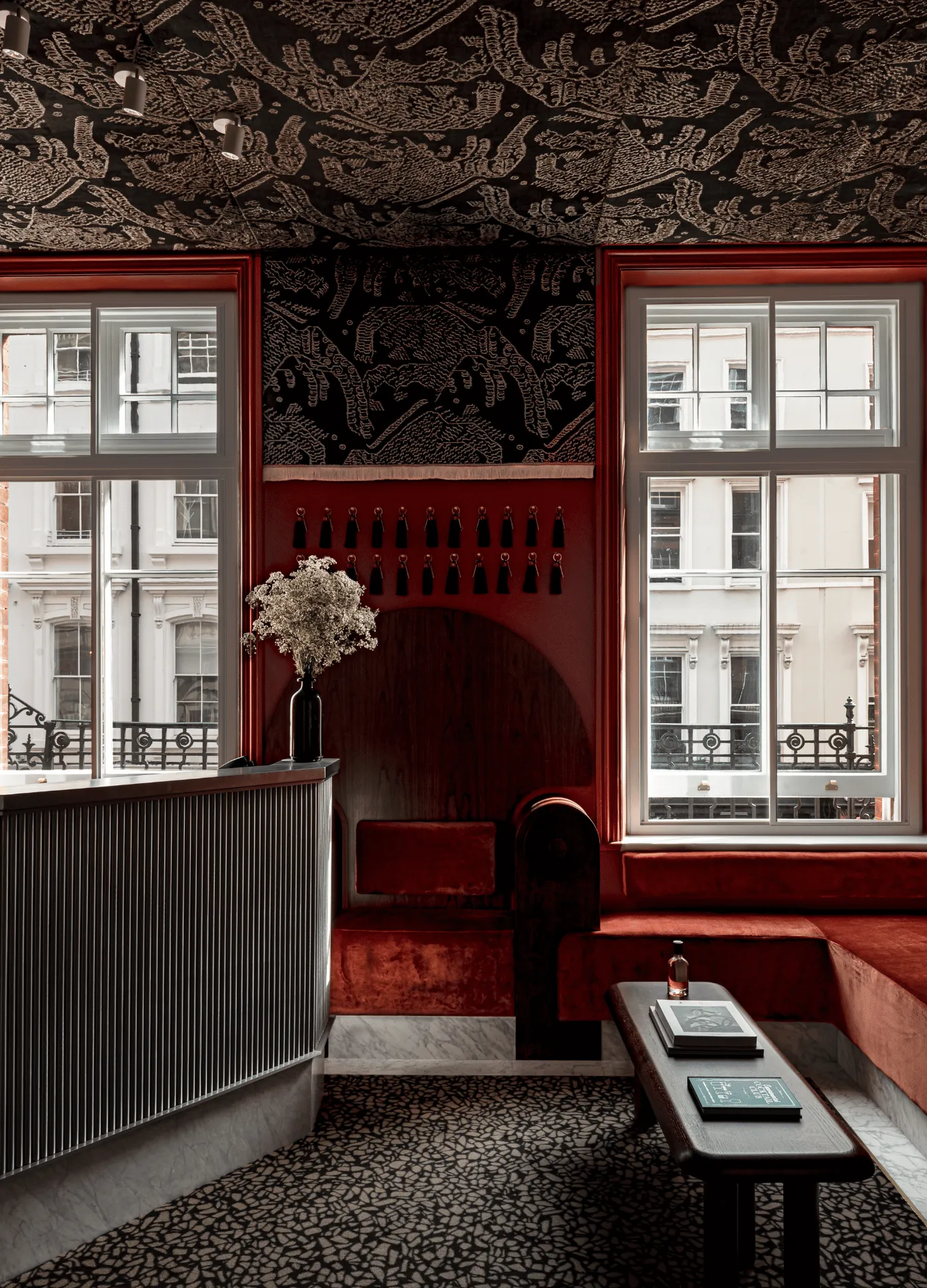 Luxurious hotel reception area with dark velvet seating, patterned ceiling, large windows, and a wooden table with books and a bottle.
