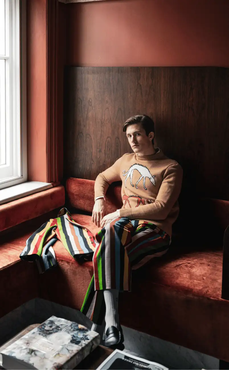 Man sitting on a rust-colored velvet couch wearing colorful striped pants and a tan sweater with a white dog design, next to a matching striped jacket and a window.