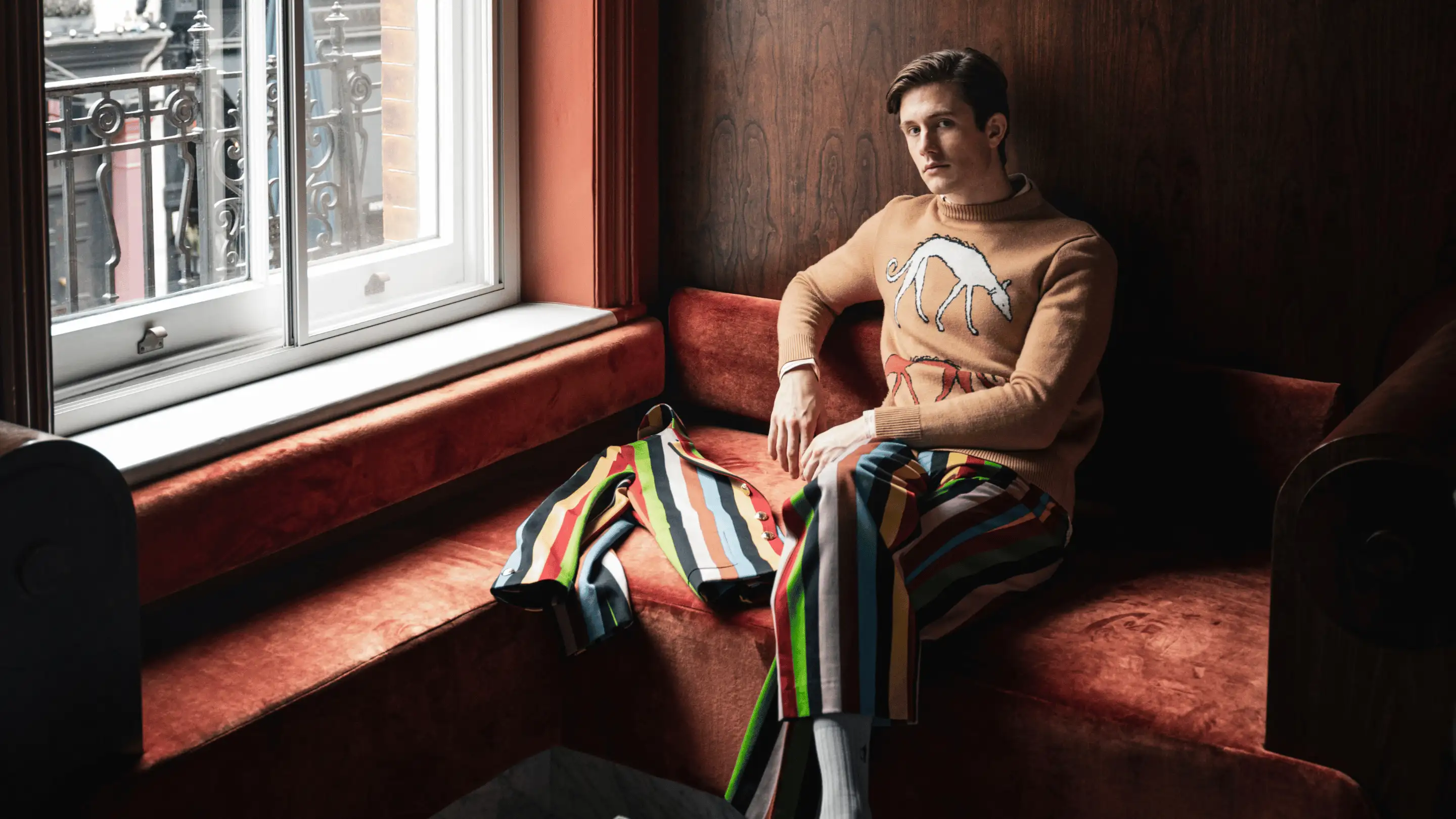Man sitting on a red velvet corner bench wearing a tan sweater with a white dog graphic and colorful striped pants, next to a colorful striped jacket.