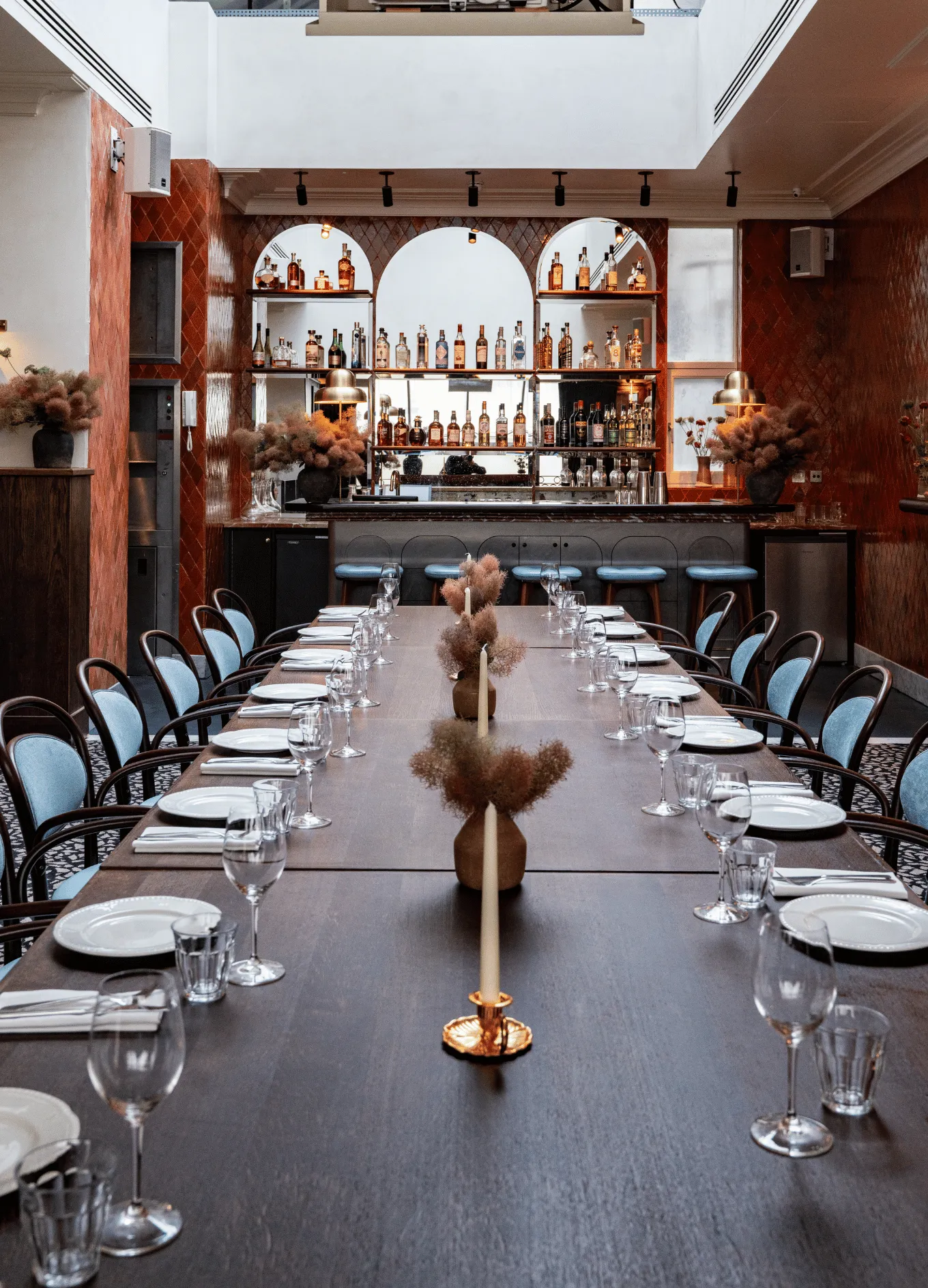 Long dining table set with plates, glasses, and napkins in front of a bar with shelves of liquor bottles and blue cushioned chairs.