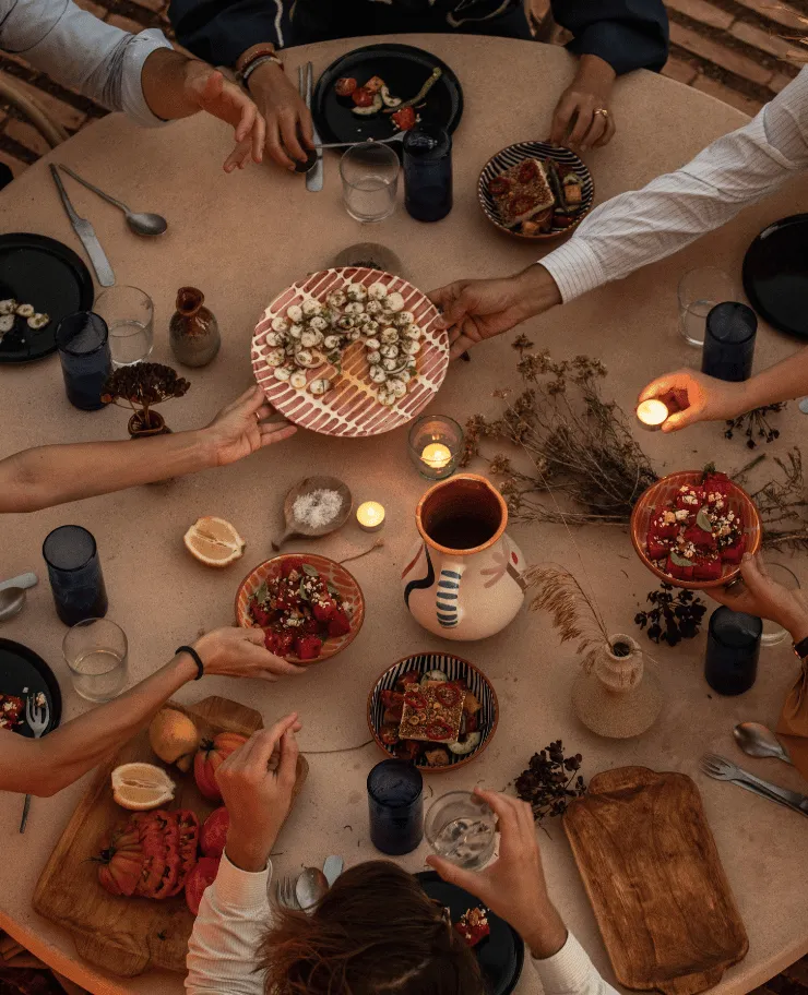 Overhead view of people sharing plates of assorted appetizers and salads around a round table with candles and rustic decor.
