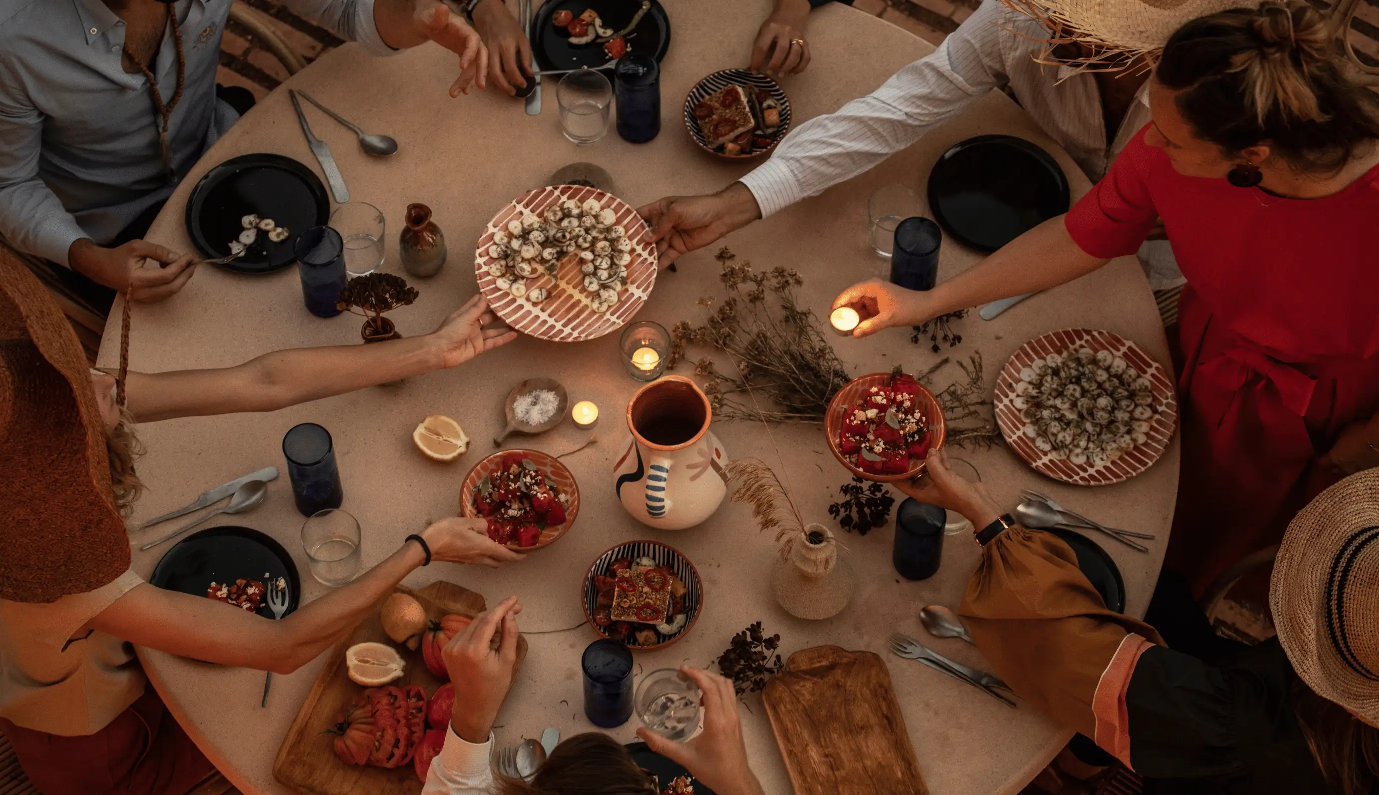 Overhead view of a group of people sharing food and lighting candles around a round table with plates of appetizers and blue drinking glasses.