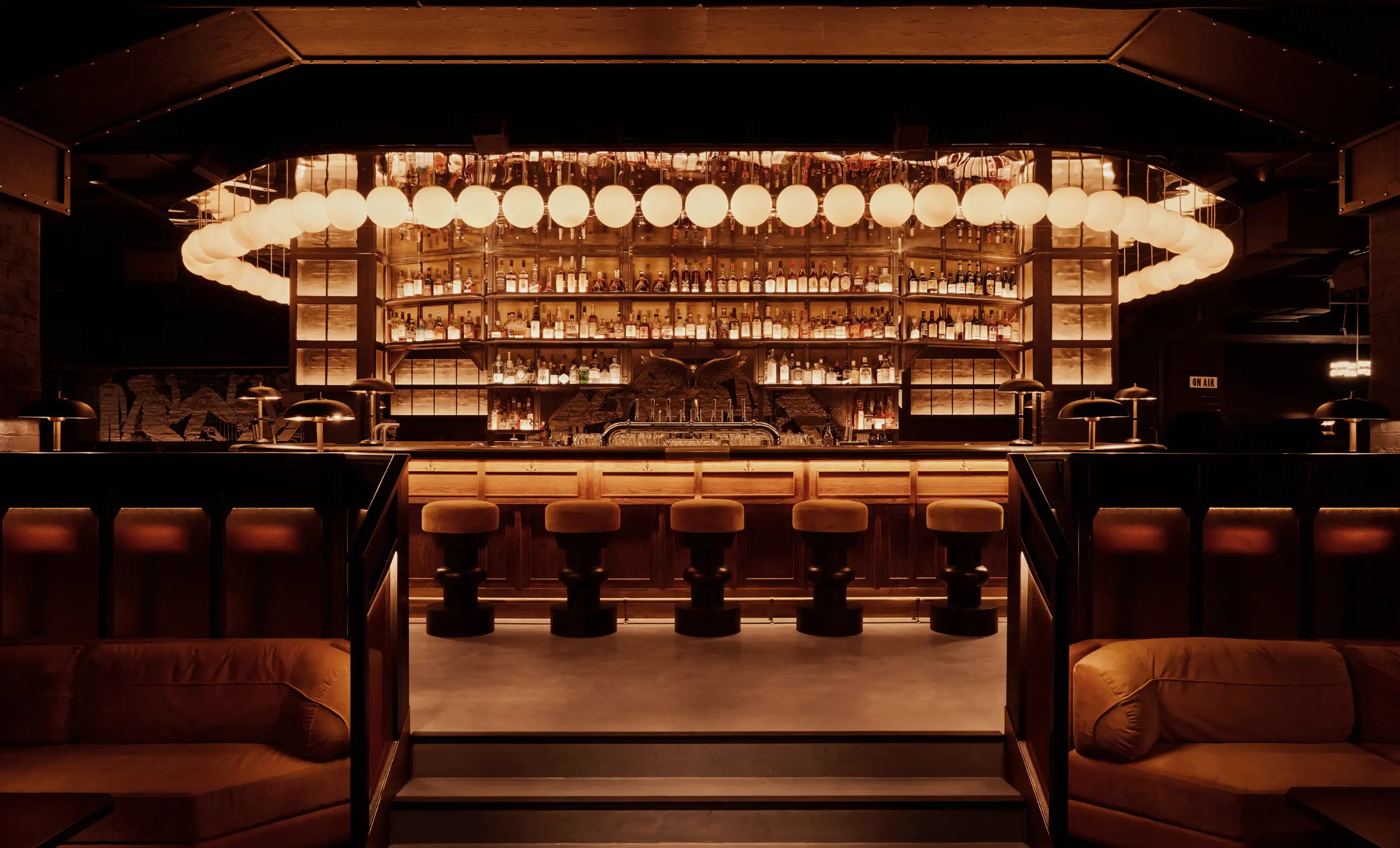 Dimly lit elegant bar with round hanging lights, wooden stools, and shelves stocked with liquor bottles.