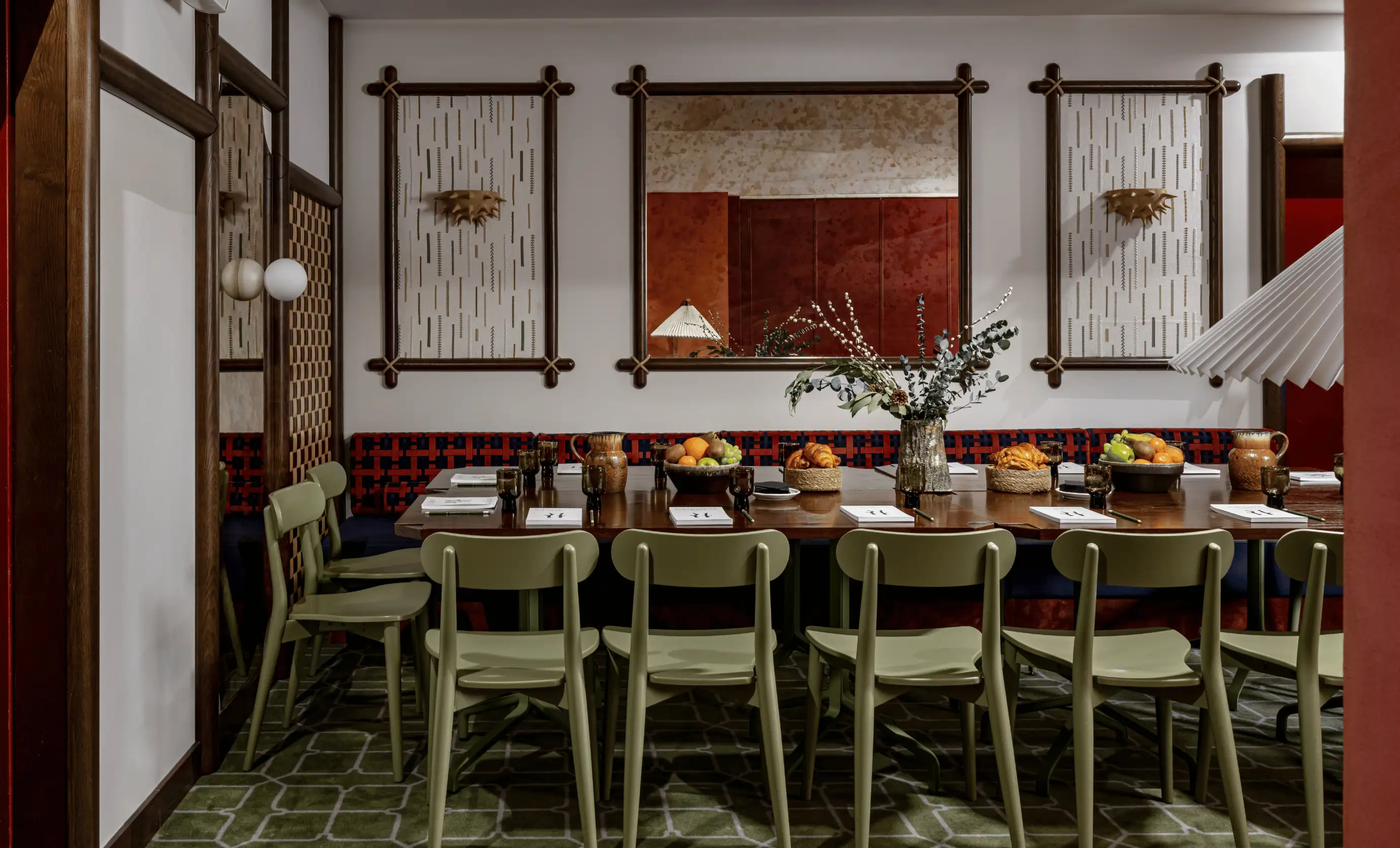 Long wooden table set for a private event with green chairs, fruit bowls, pastries, and vase with greenery in a warmly decorated room.