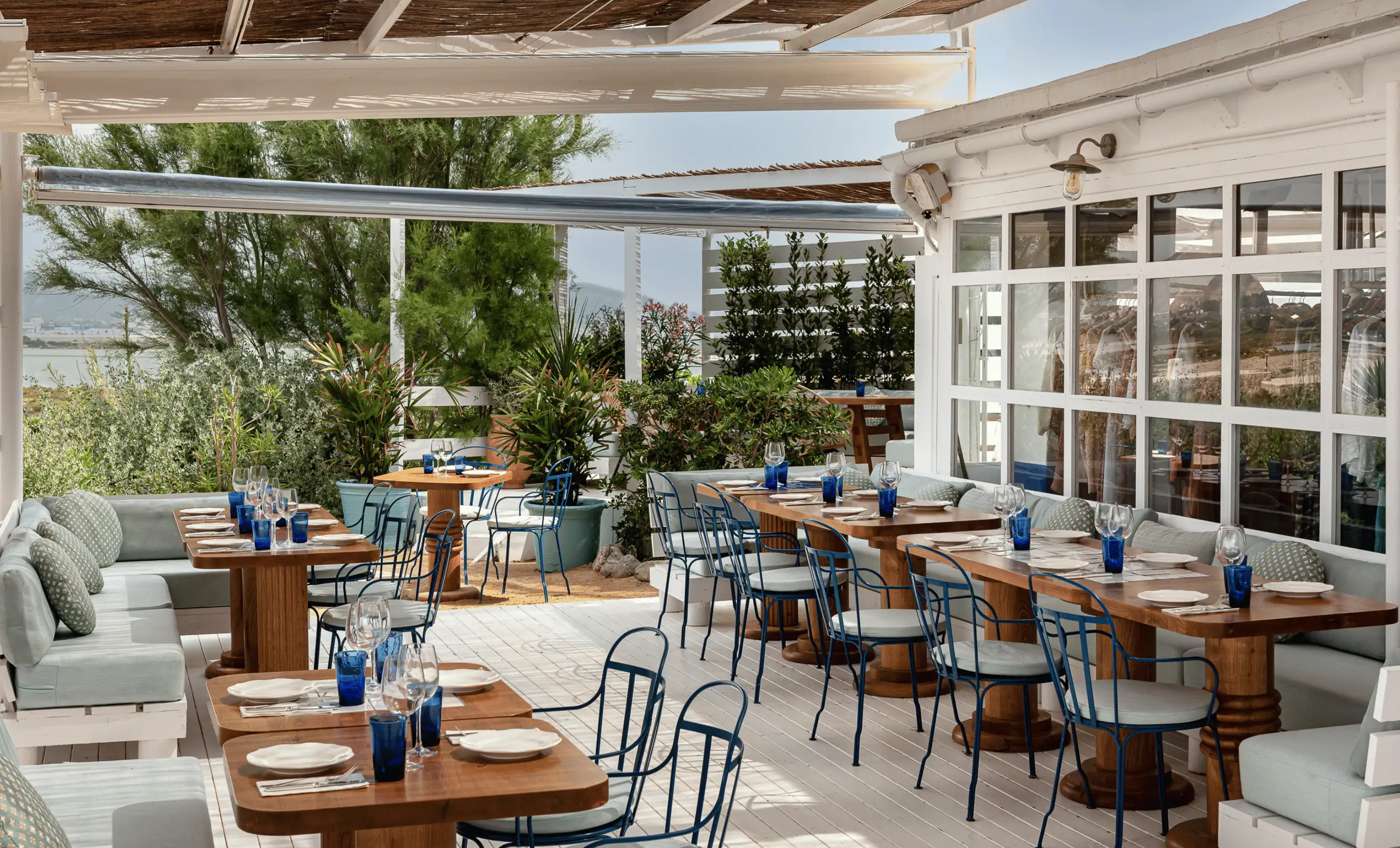 Outdoor dining area with wooden tables, blue metal chairs, cushioned benches, and plants along a white deck.