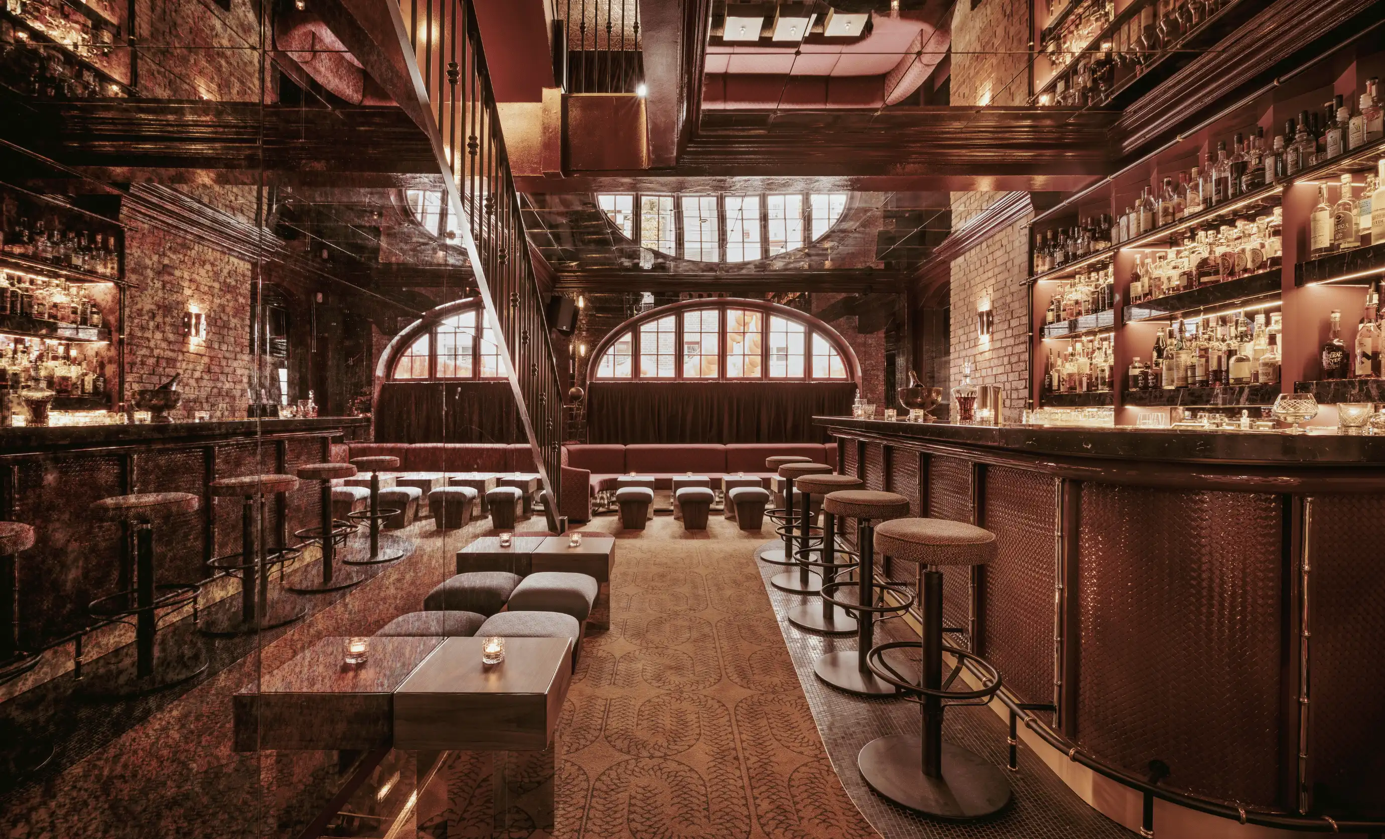 Elegant hotel bar with high stools, wooden tables, mirrored wall, exposed brick, and shelves stocked with bottles.